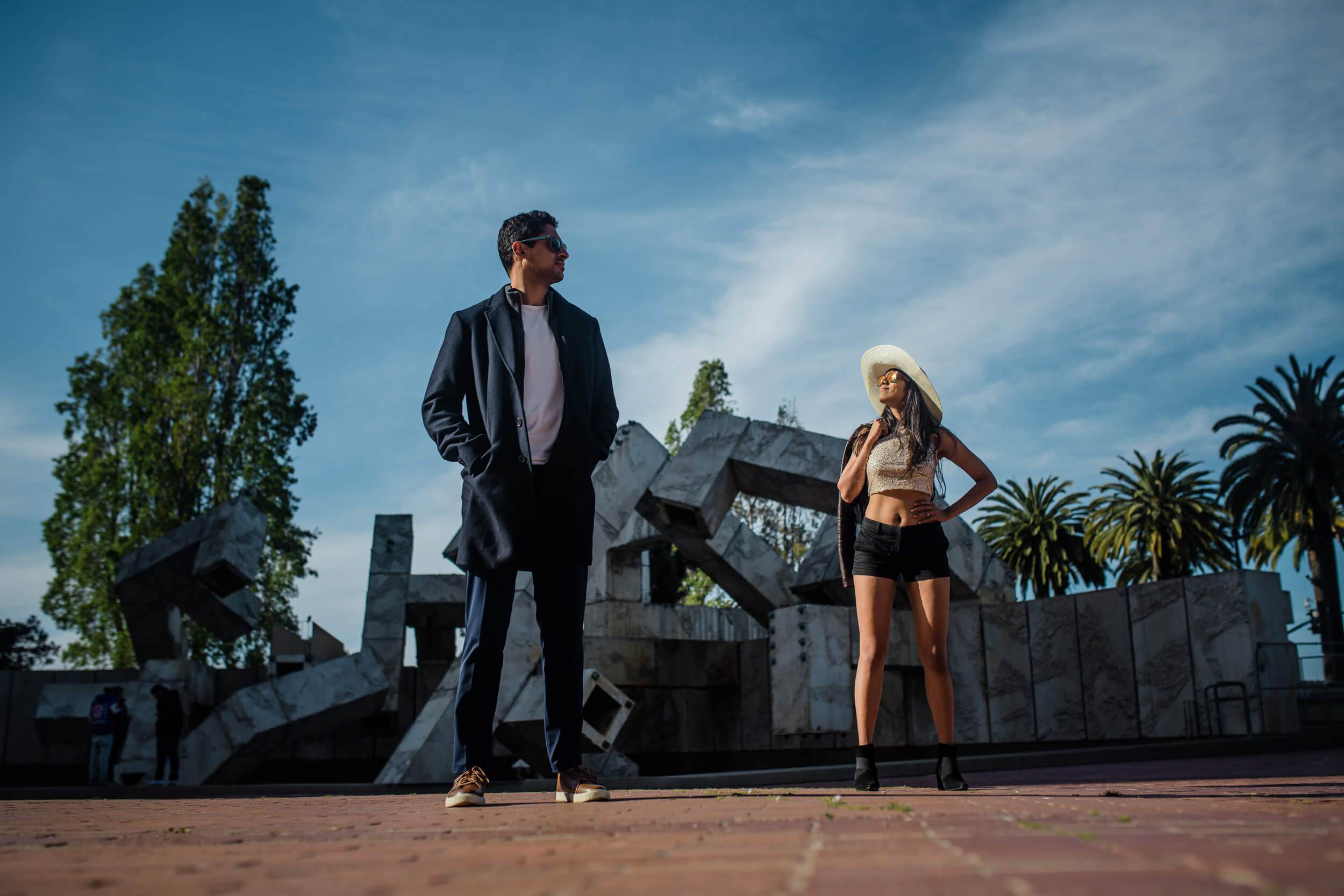 Anu and Raghu stand apart on a California plaza with palm trees and sculptural architecture under blue sky