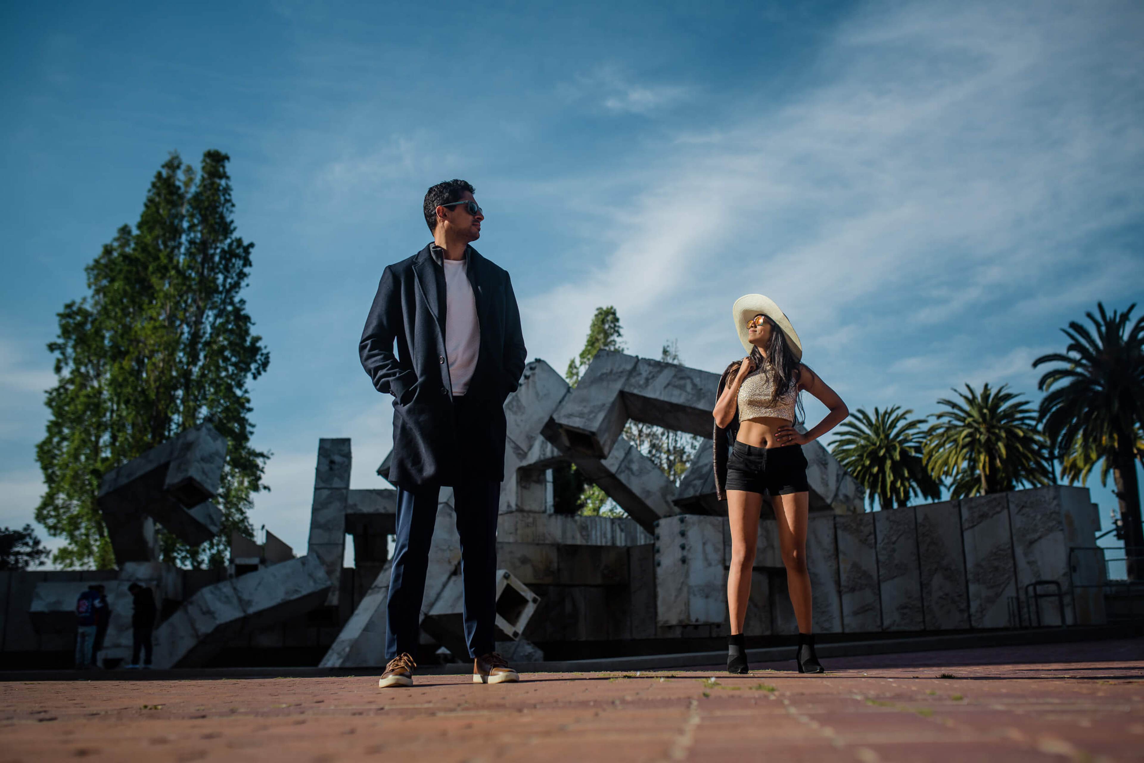 Anu and Raghu stand apart on a California plaza with palm trees and sculptural architecture under blue sky