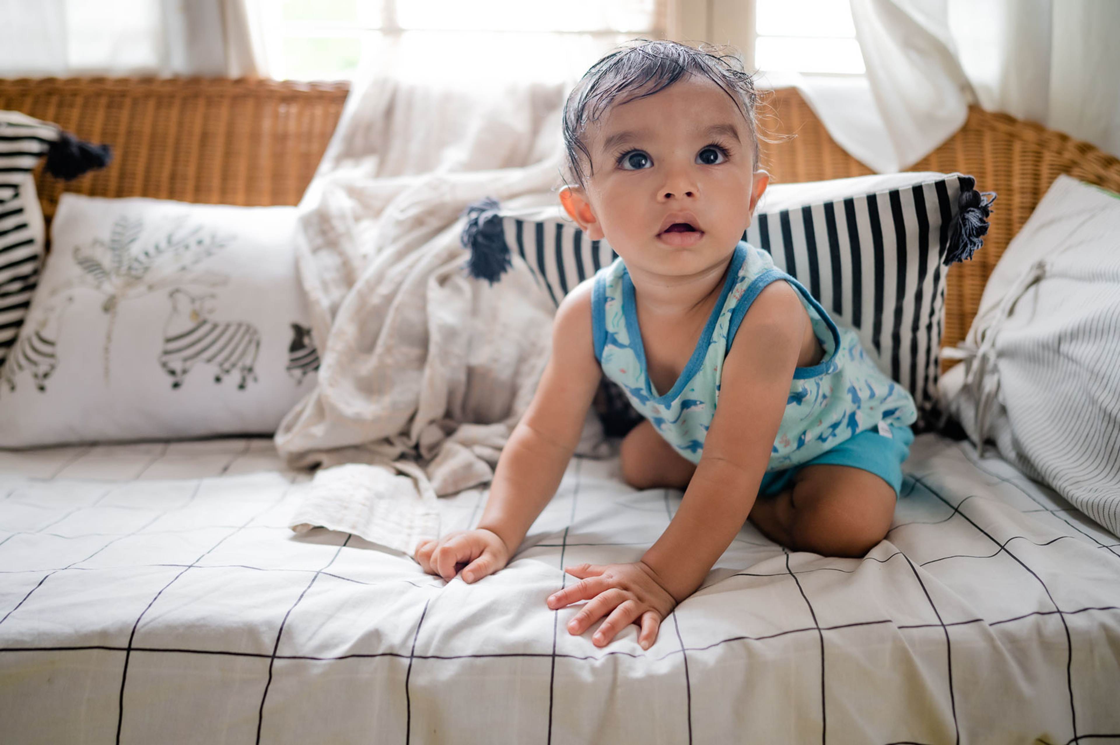 Toddler in blue outfit crawling on bed with patterned pillows and bright window light