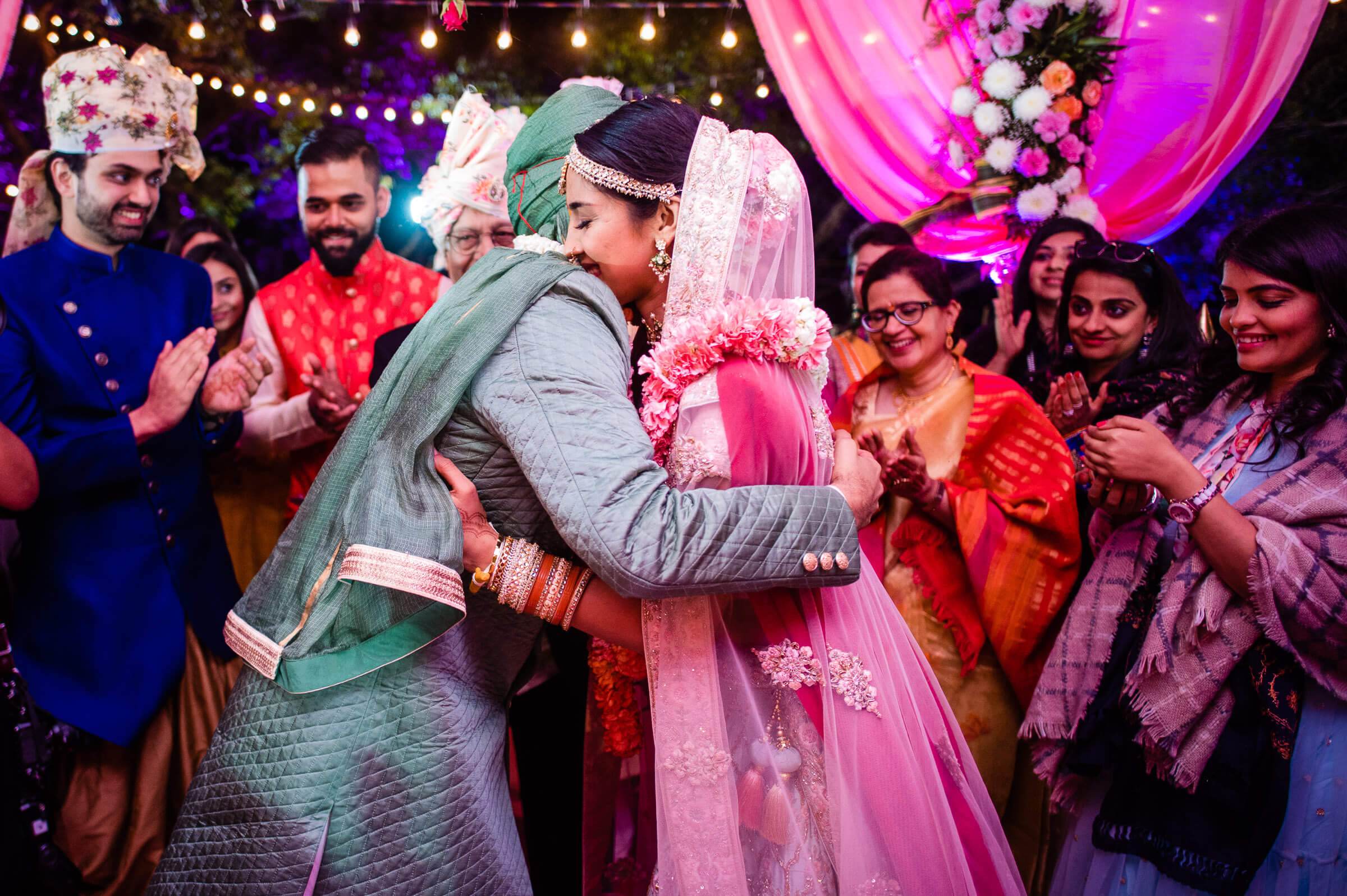 Shreya and Yash embrace during vibrant pink-lit reception at Mahabaleshwar, surrounded by smiling guests in traditional attire