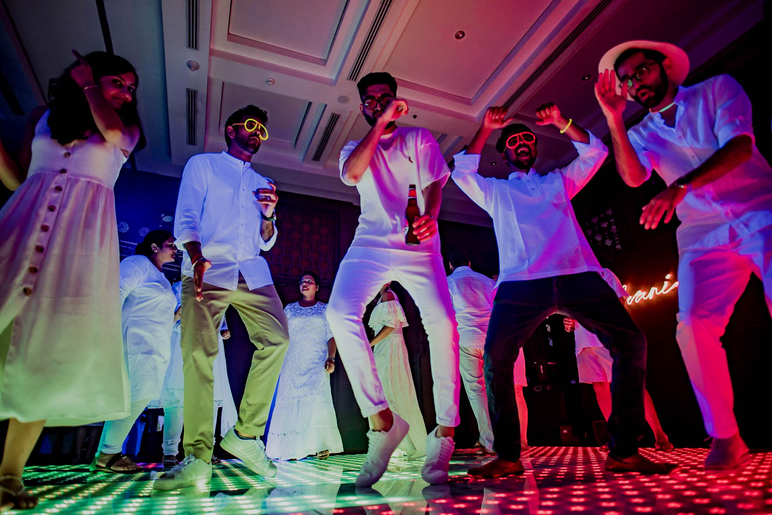 Avani & Srikant's wedding guests dancing under vibrant pink and blue neon lights on illuminated dance floor