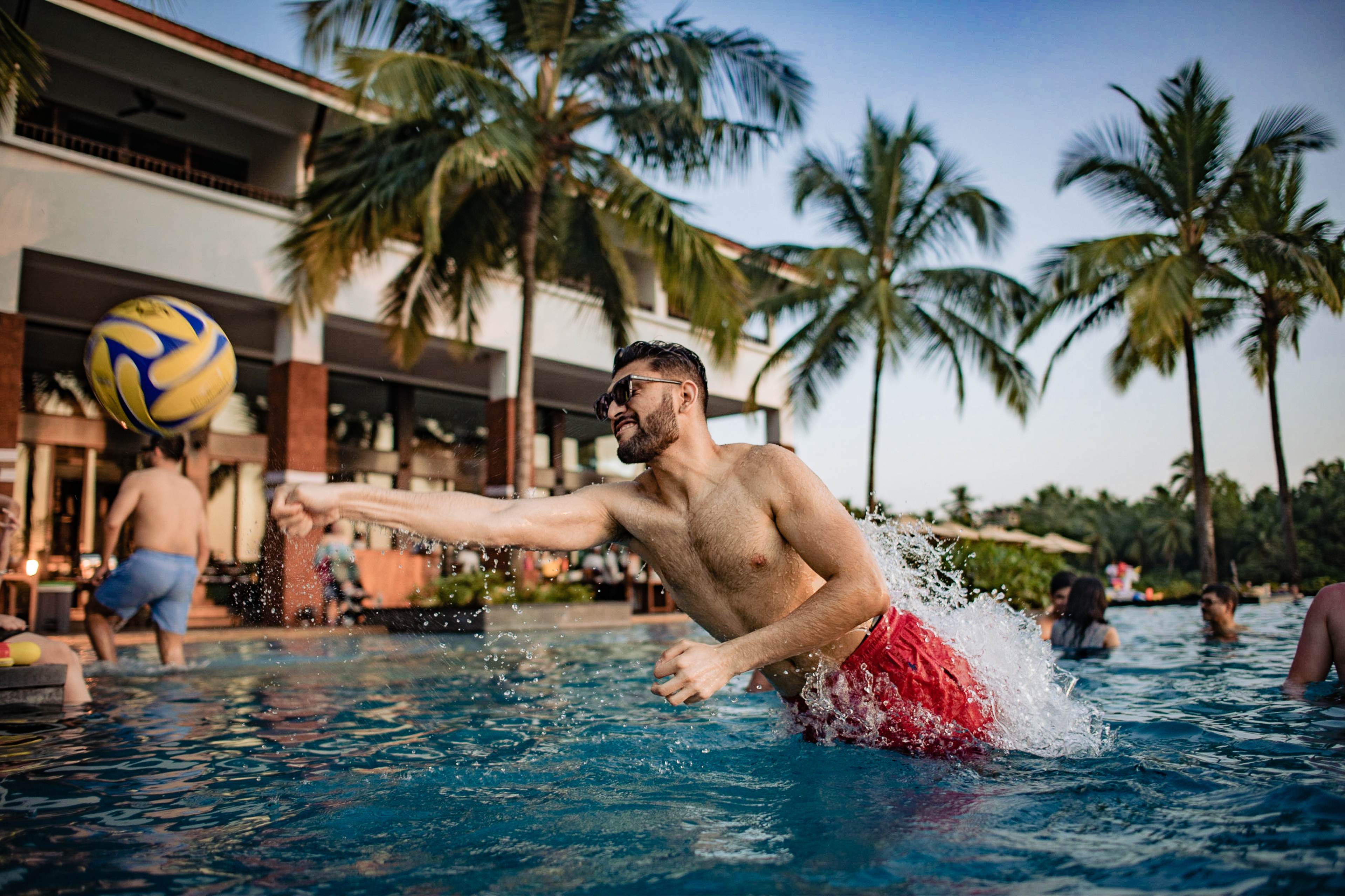 Srikant playing volleyball in a poolside game in Goa, surrounded by palm trees and tropical architecture