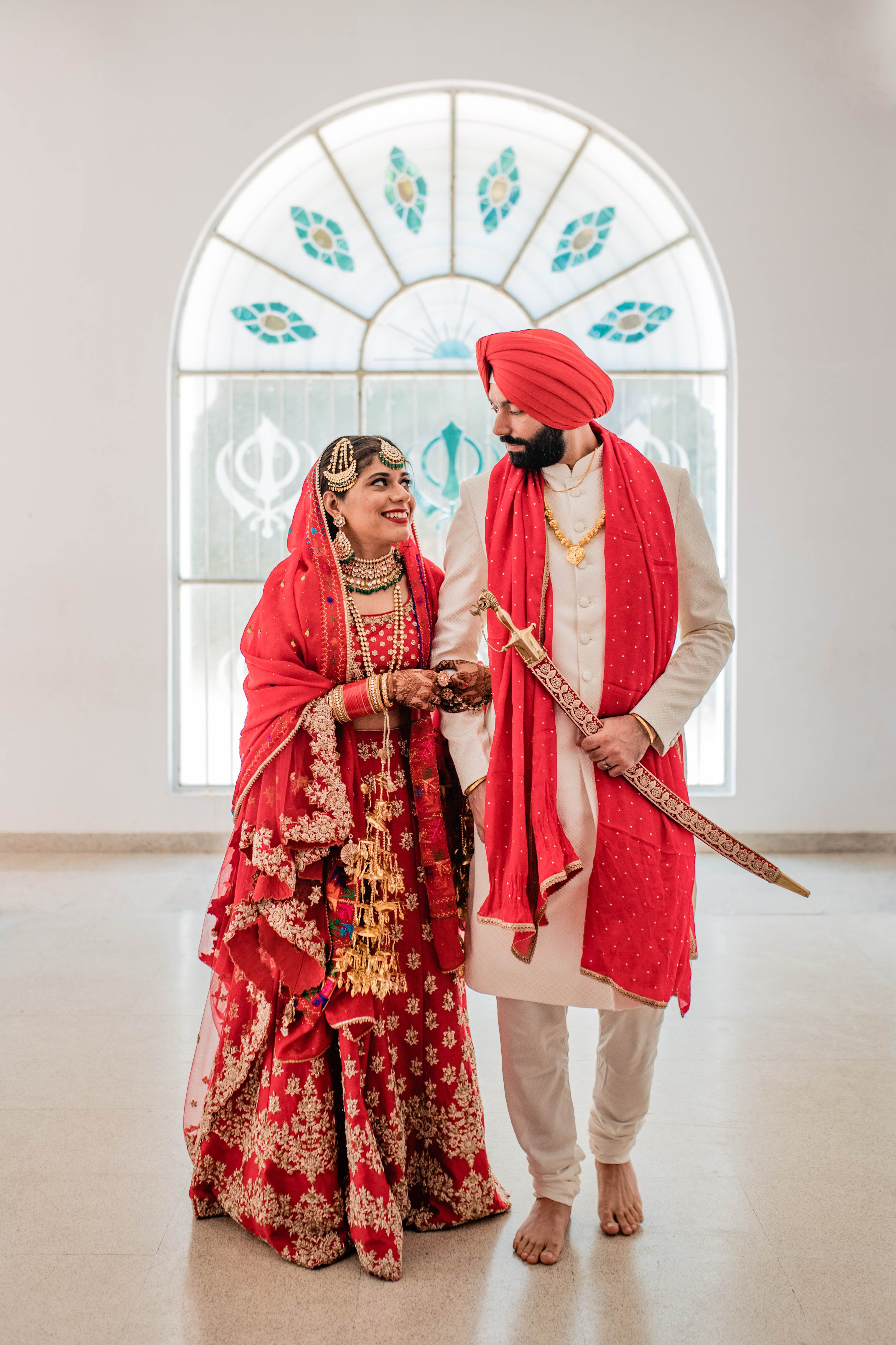 Couple in vibrant red traditional wedding attire hold hands beneath an arched window with blue patterned glass