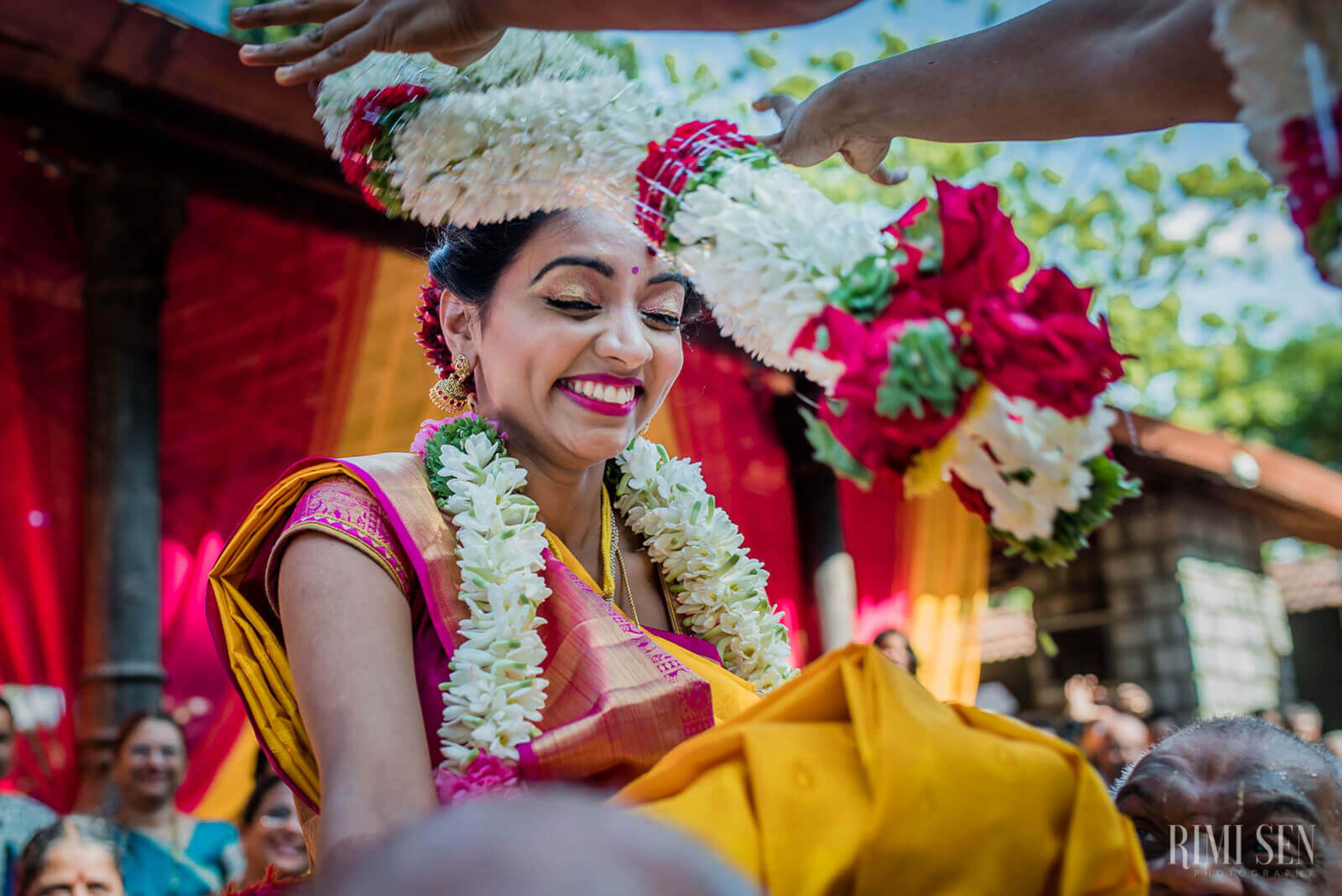 Pooja smiling in pink wedding attire with white flower garlands, hands adjusting her headpiece, vibrant red and yellow backdrop
