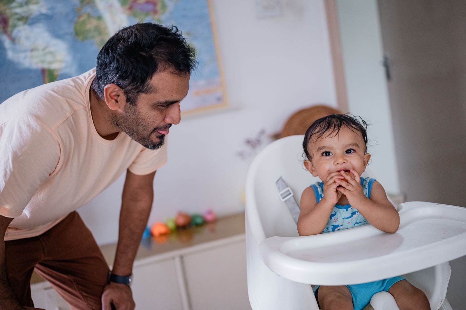 Man in beige shirt leans toward toddler in high chair, bright playful moment indoors