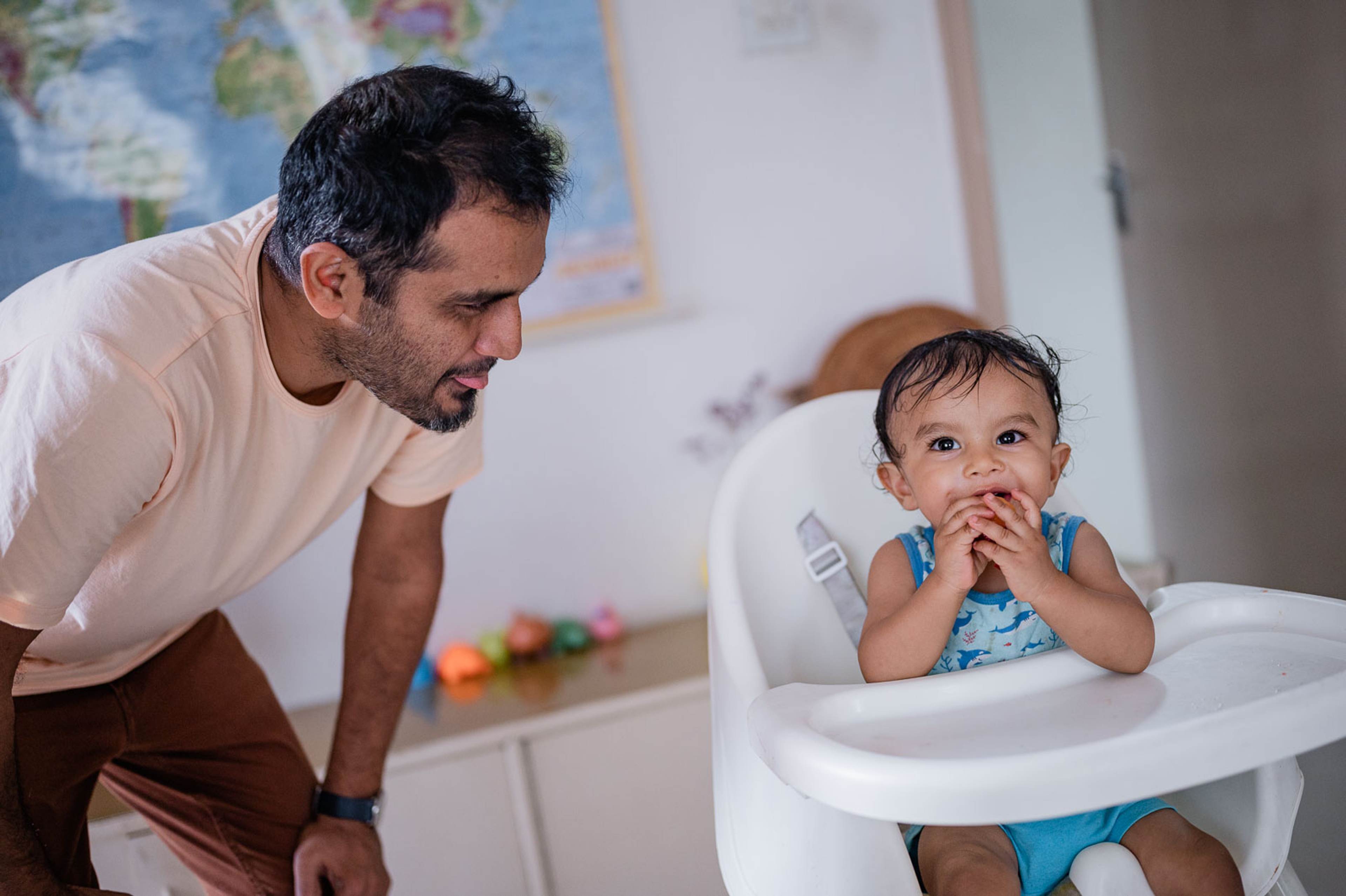 Man in beige shirt leans toward toddler in high chair, bright playful moment indoors