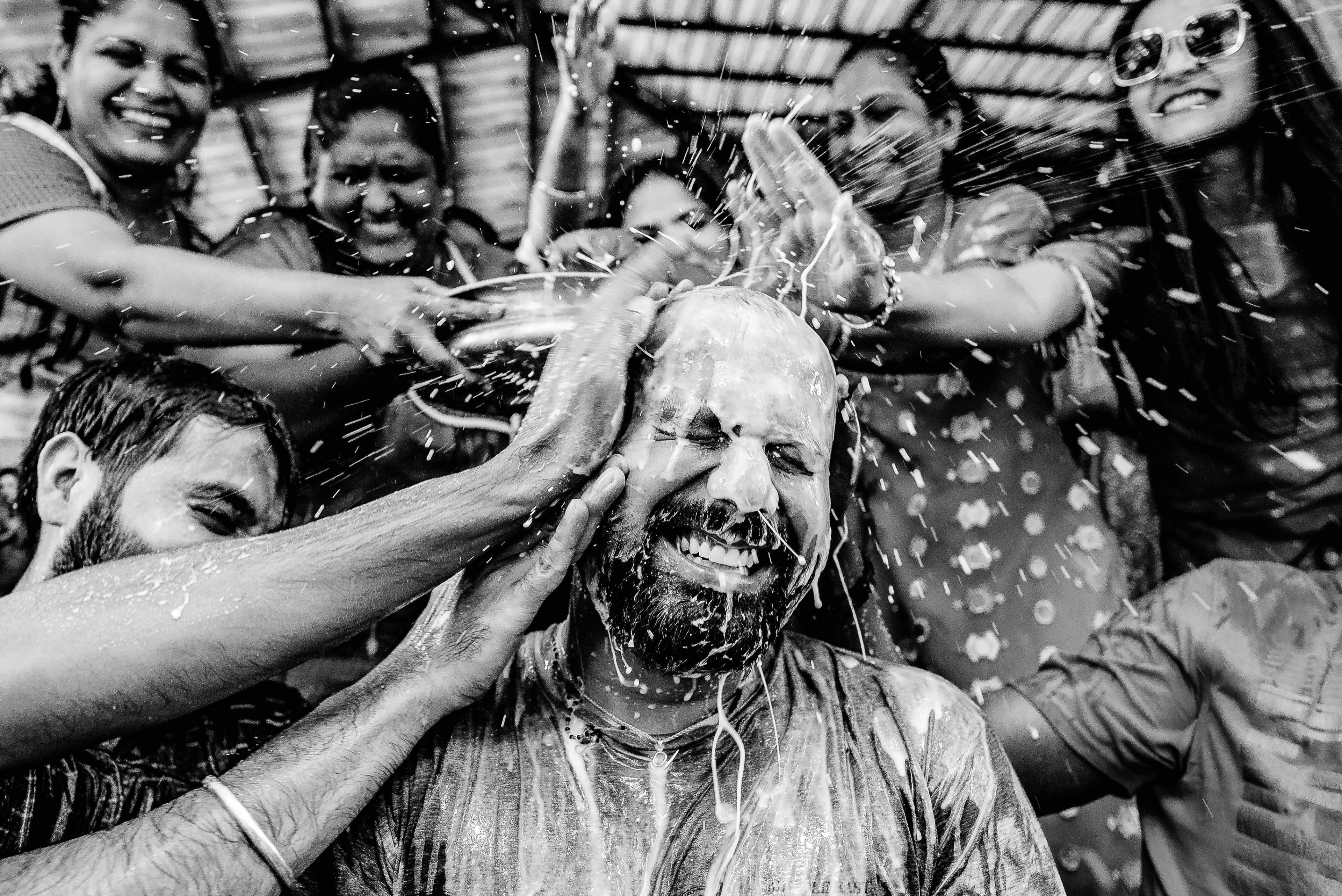 Groom smiling as wedding guests pour water over his head in celebratory Holi ritual