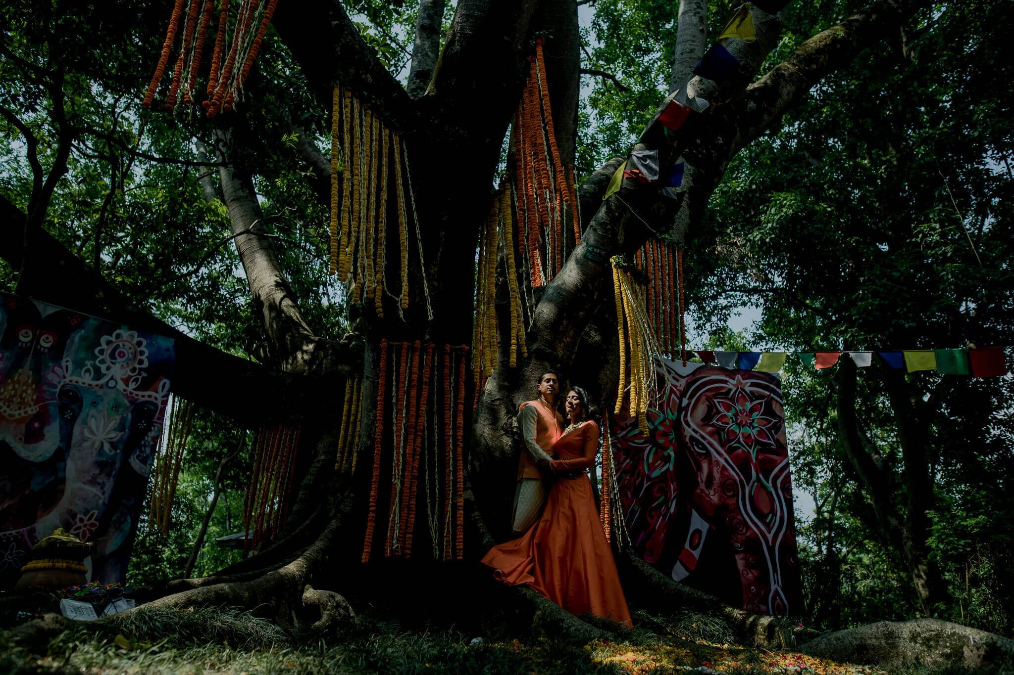 Anu and Raghu embrace under a massive ancient tree draped with golden strings, Nepal