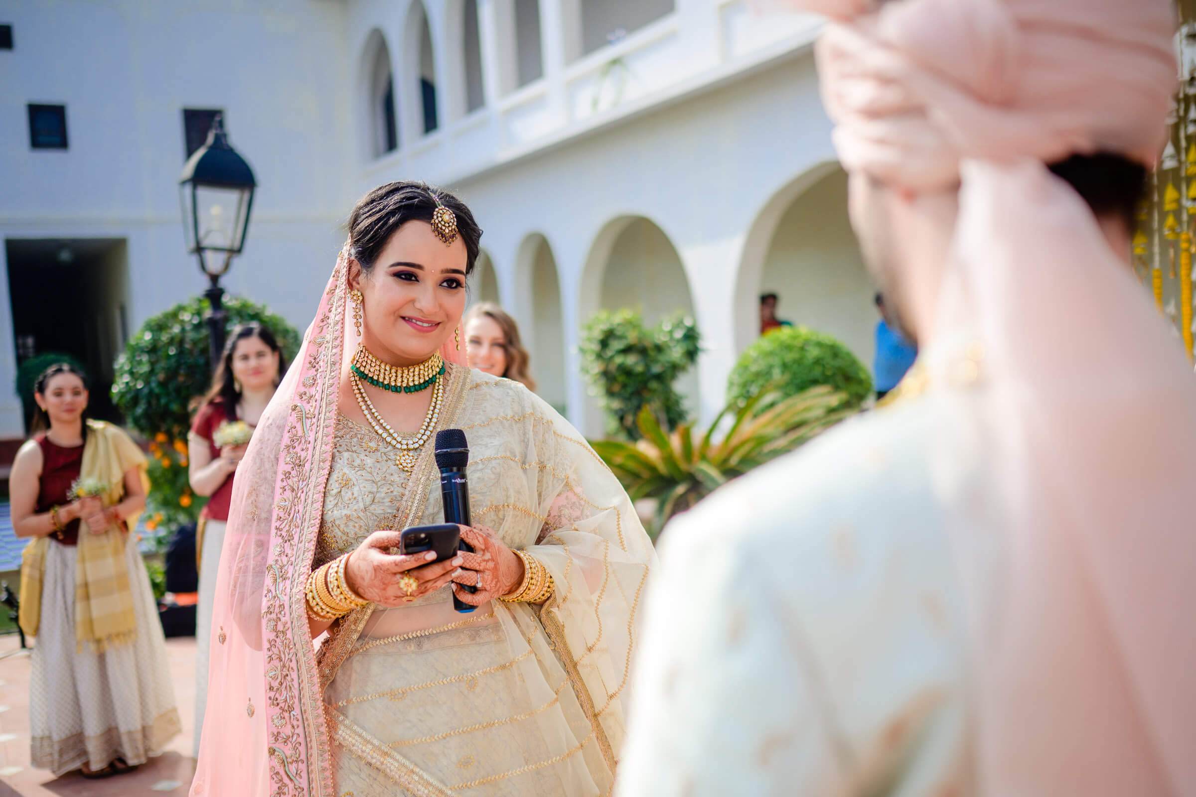 Maitrayee in ornate cream lehenga and pink dupatta holding microphone, speaking at Rajasthan wedding celebration