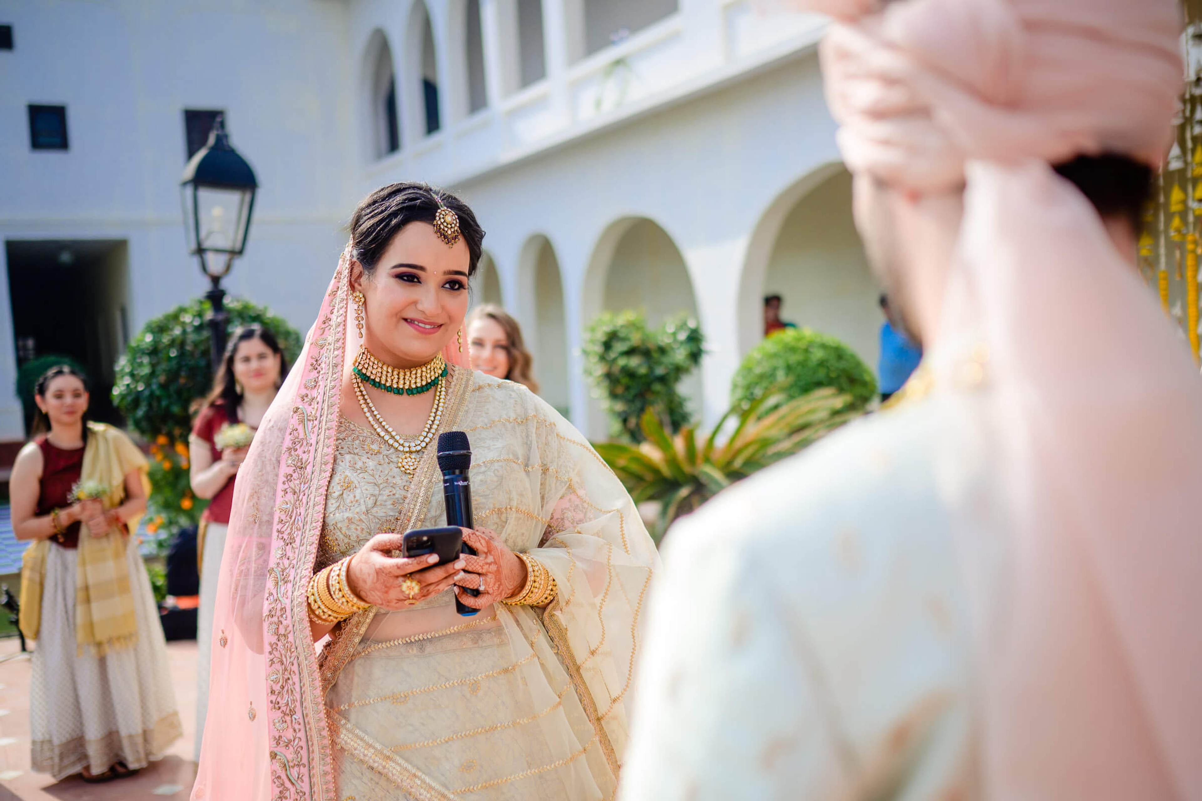 Maitrayee in ornate cream lehenga and pink dupatta holding microphone, speaking at Rajasthan wedding celebration