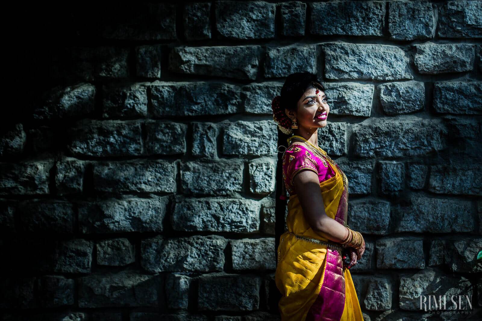 Pooja in yellow and pink saree smiling against textured stone wall, Bangalore