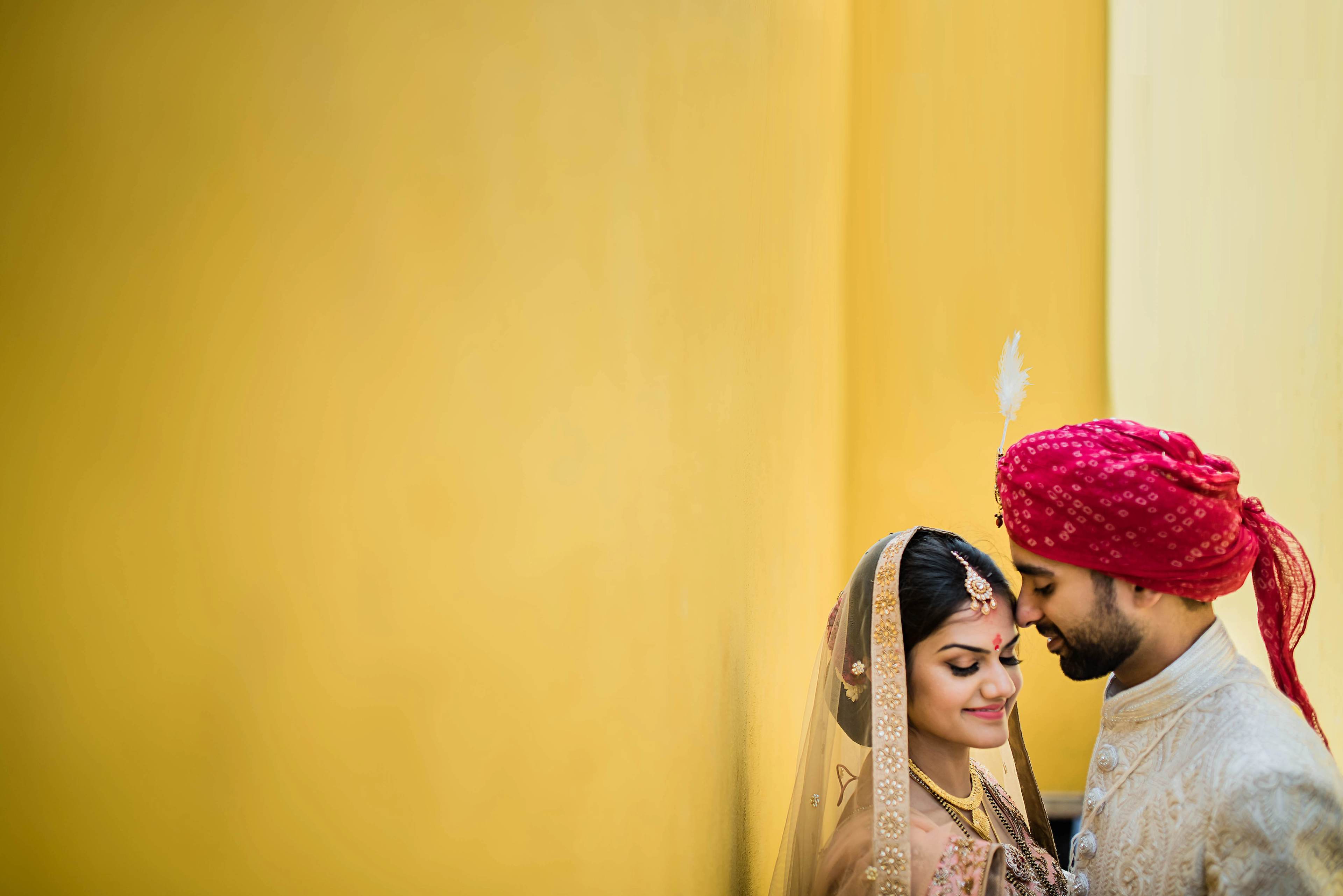 Couple in traditional wedding attire embracing against bright yellow backdrop, intimate moment