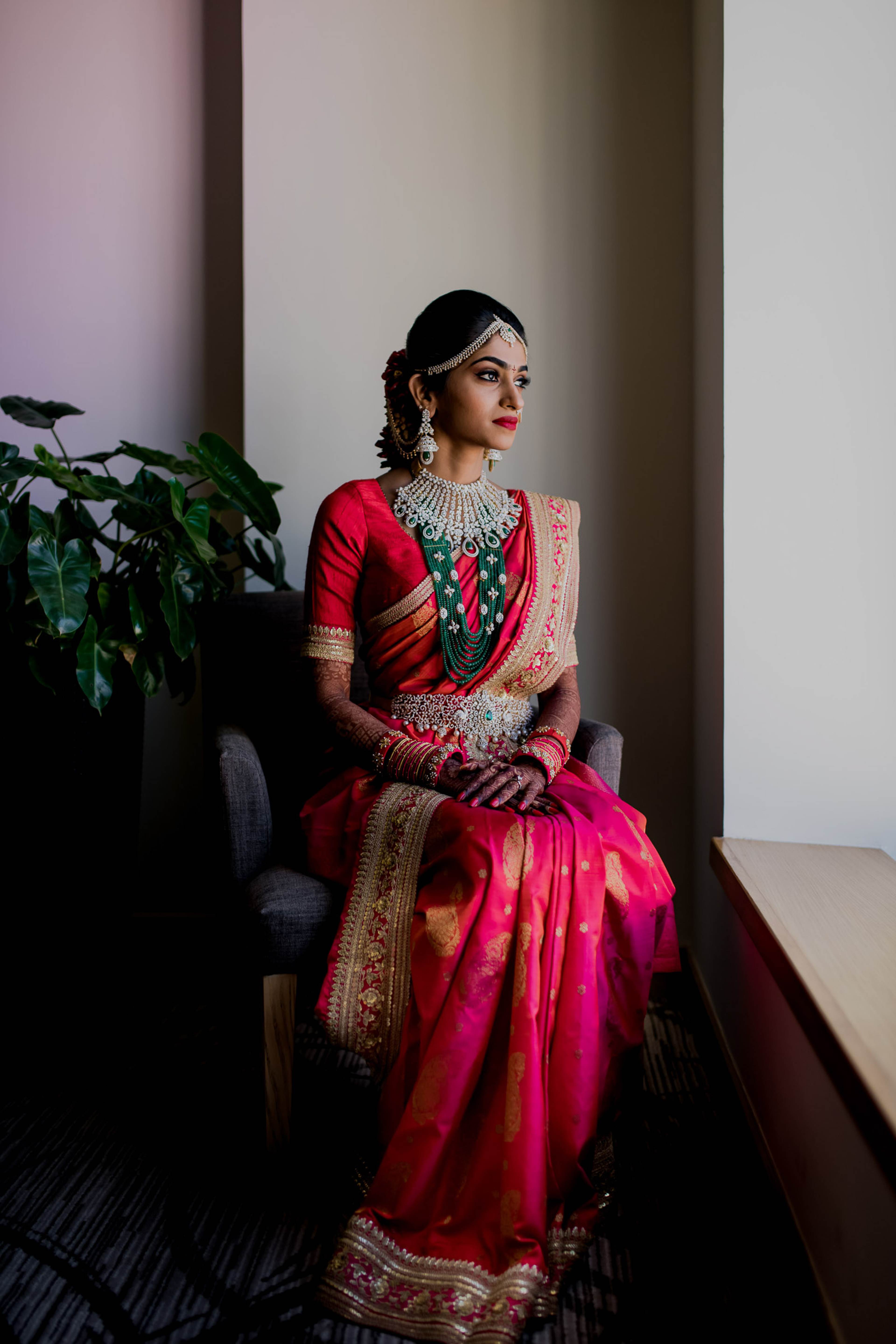 Woman in red embroidered lehenga and jeweled necklace seated by plant, gazing aside