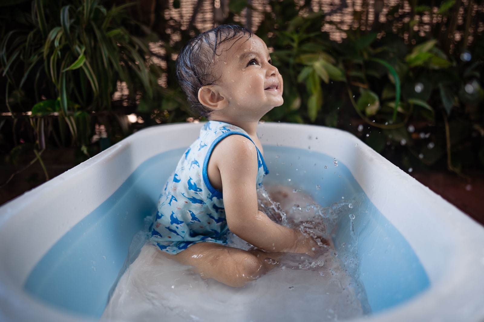 Child in blue swimsuit bathing in white tub with water, surrounded by greenery