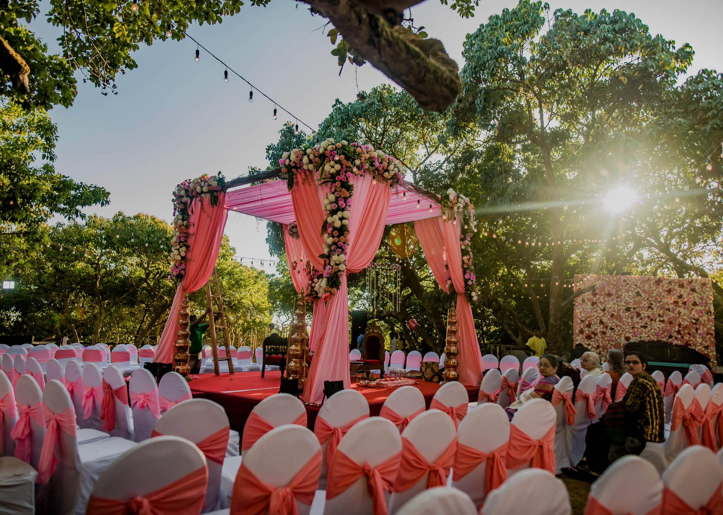 Shreya and Yash's wedding mandap with pink drapes and floral garland, surrounded by white chairs with coral sashes in Mahabaleshwar garden