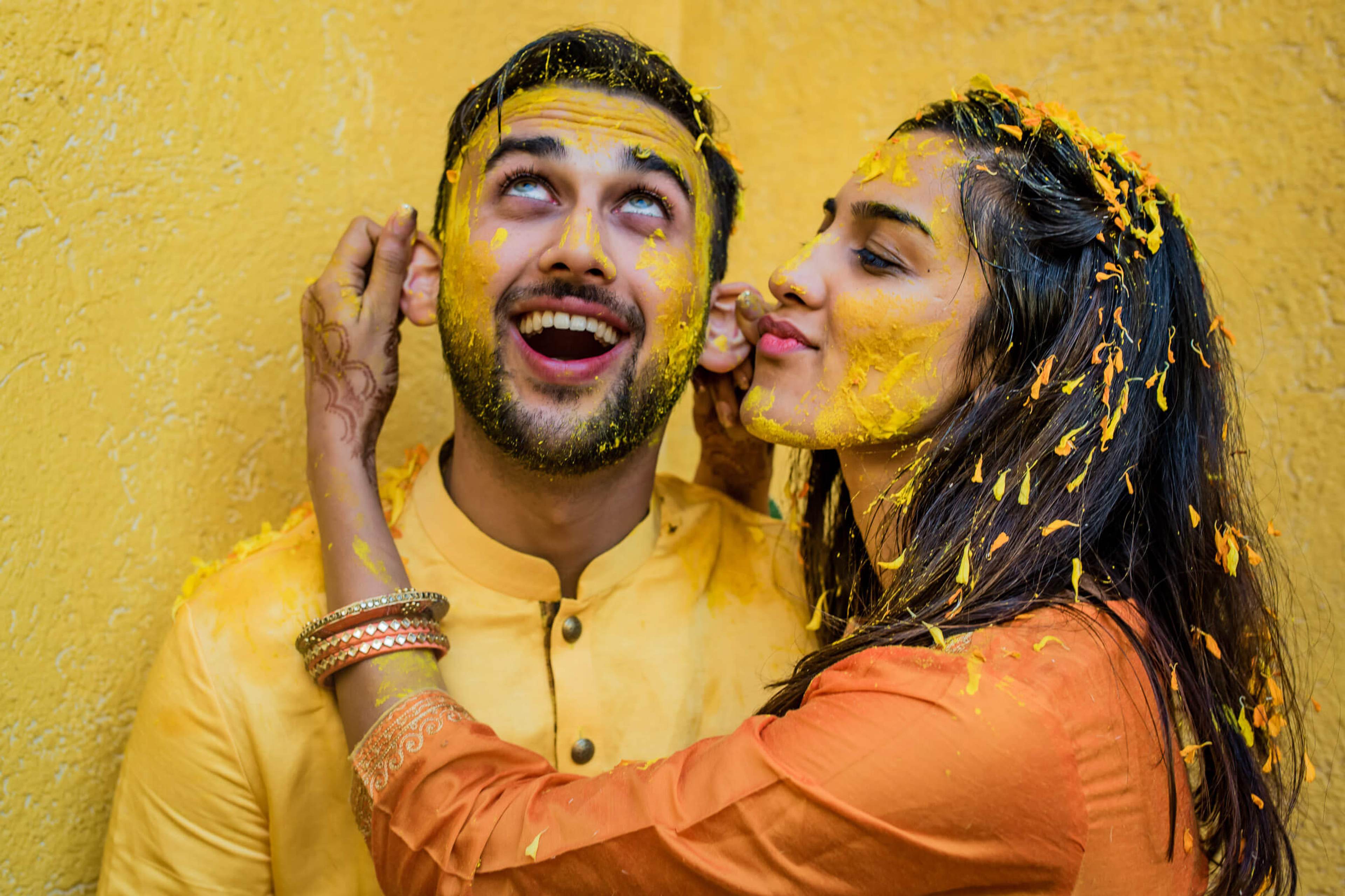Shreya and Yash laughing together with gold paint against bright yellow wall