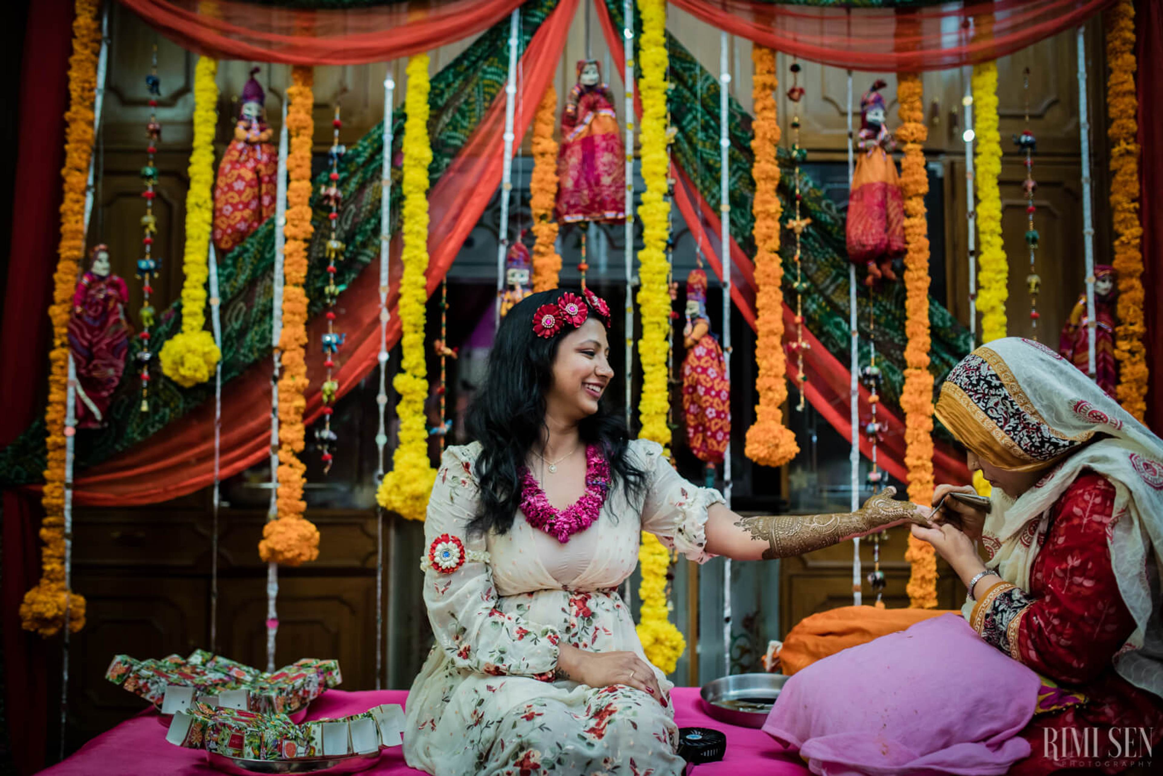 Nivetha in white floral attire, adorned with pink garland, sits amid vibrant hanging flowers and red fabric draping at Bangalore wedding celebration