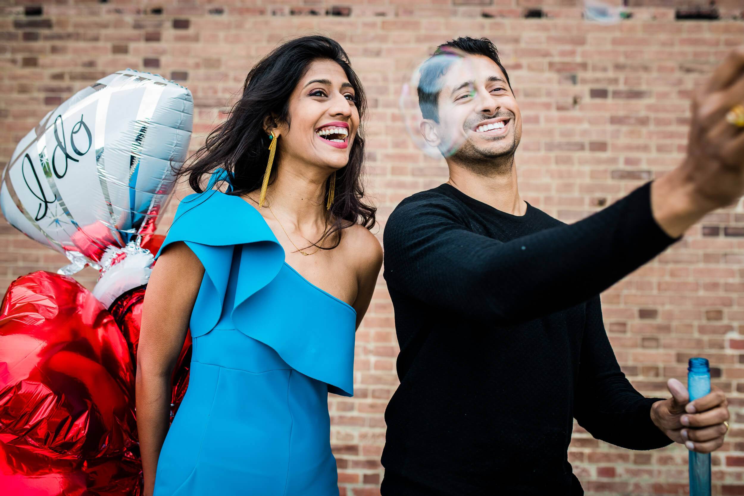 Anu and Raghu hold festive balloons against a brick wall, smiling and looking skyward in California