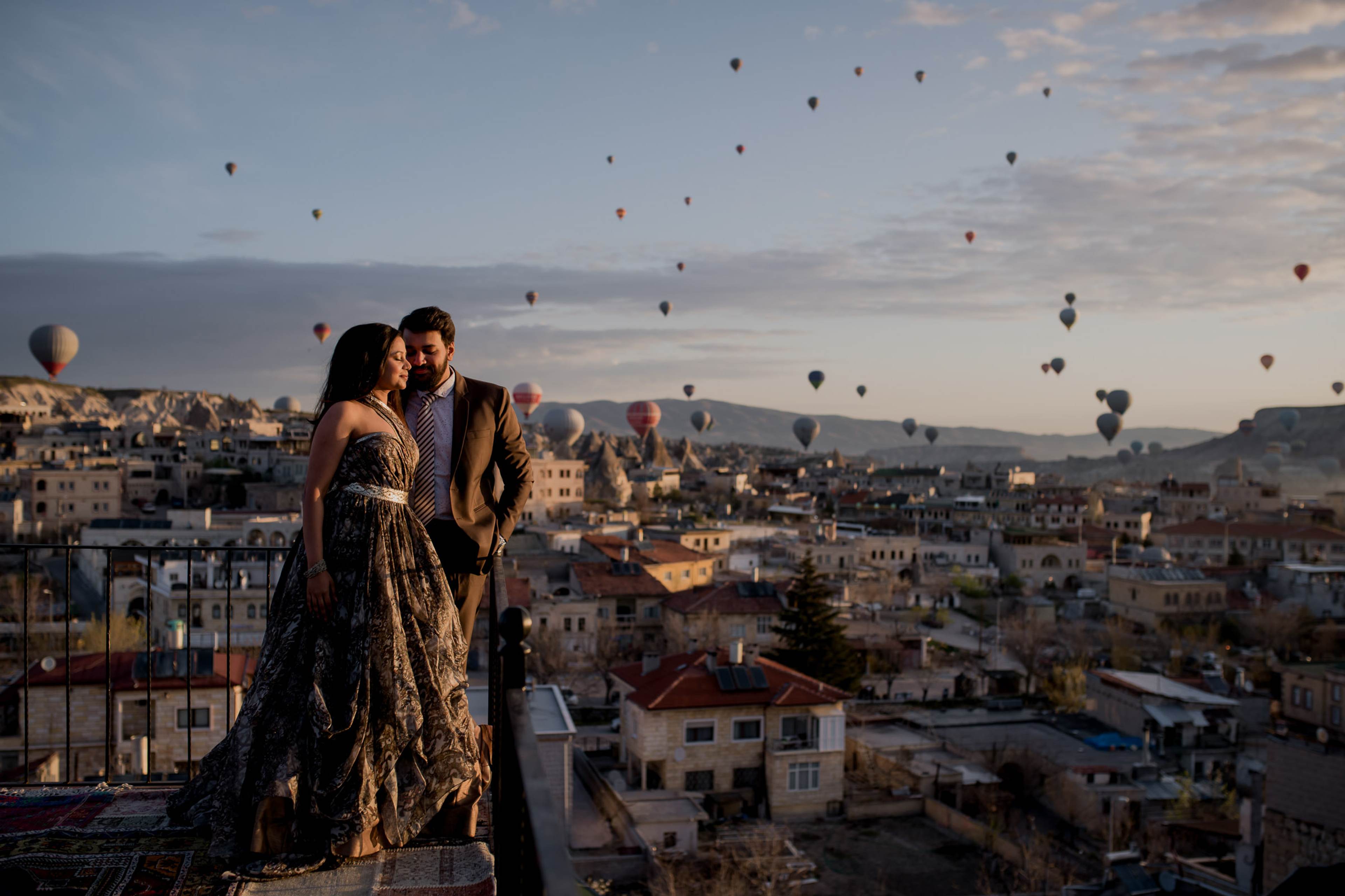 Couple embracing on rooftop at sunrise with hot air balloons floating above Cappadocia's terracotta buildings