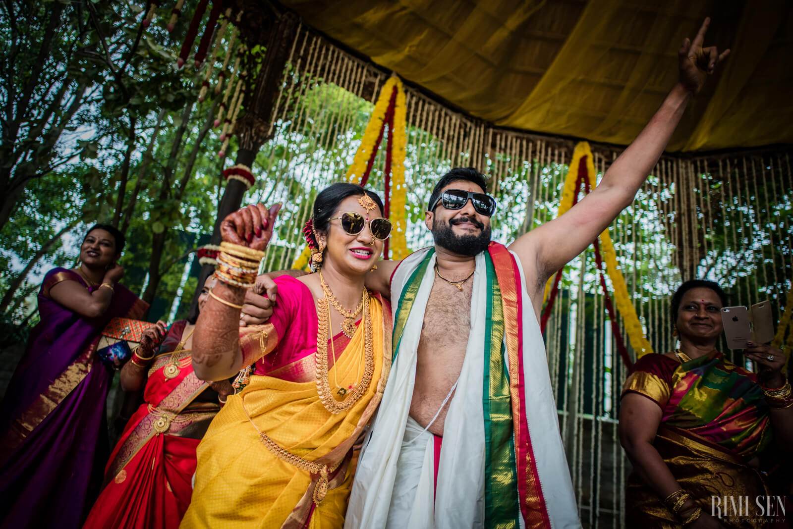 Nivetha and Sridhar celebrate under a yellow canopy, wearing traditional attire and sunglasses, surrounded by smiling guests in Bangalore