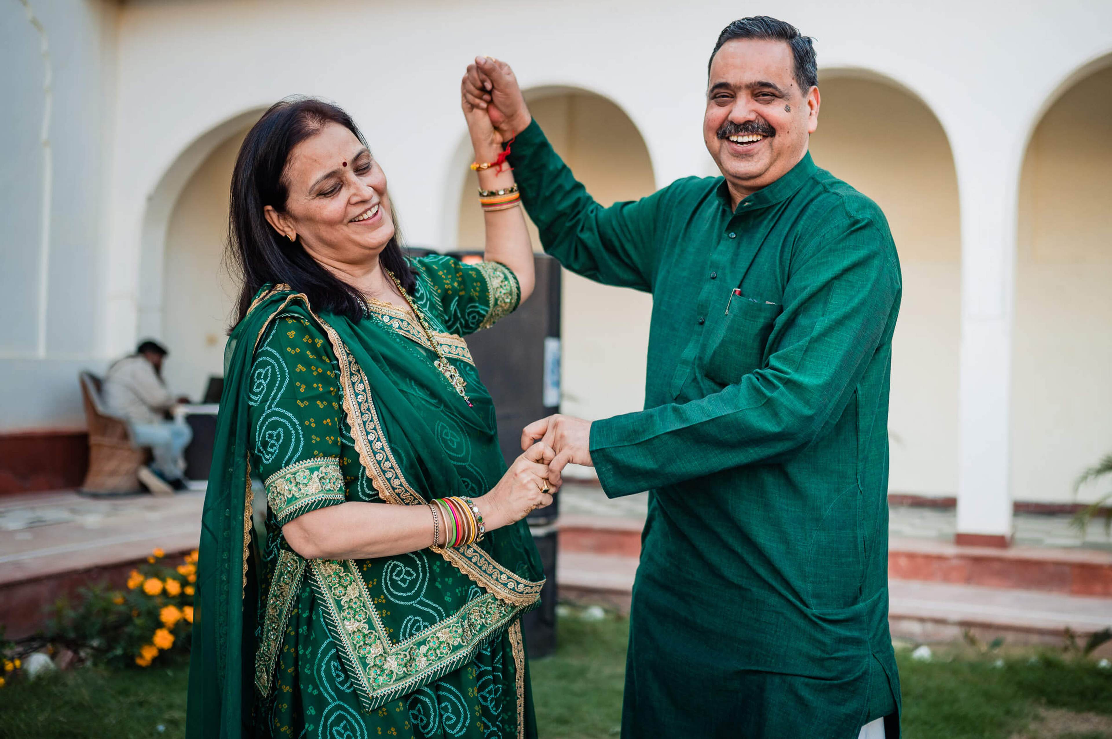 Maitrayee and Eric celebrate in matching emerald traditional attire, hands raised joyfully before arched architecture in Rajasthan