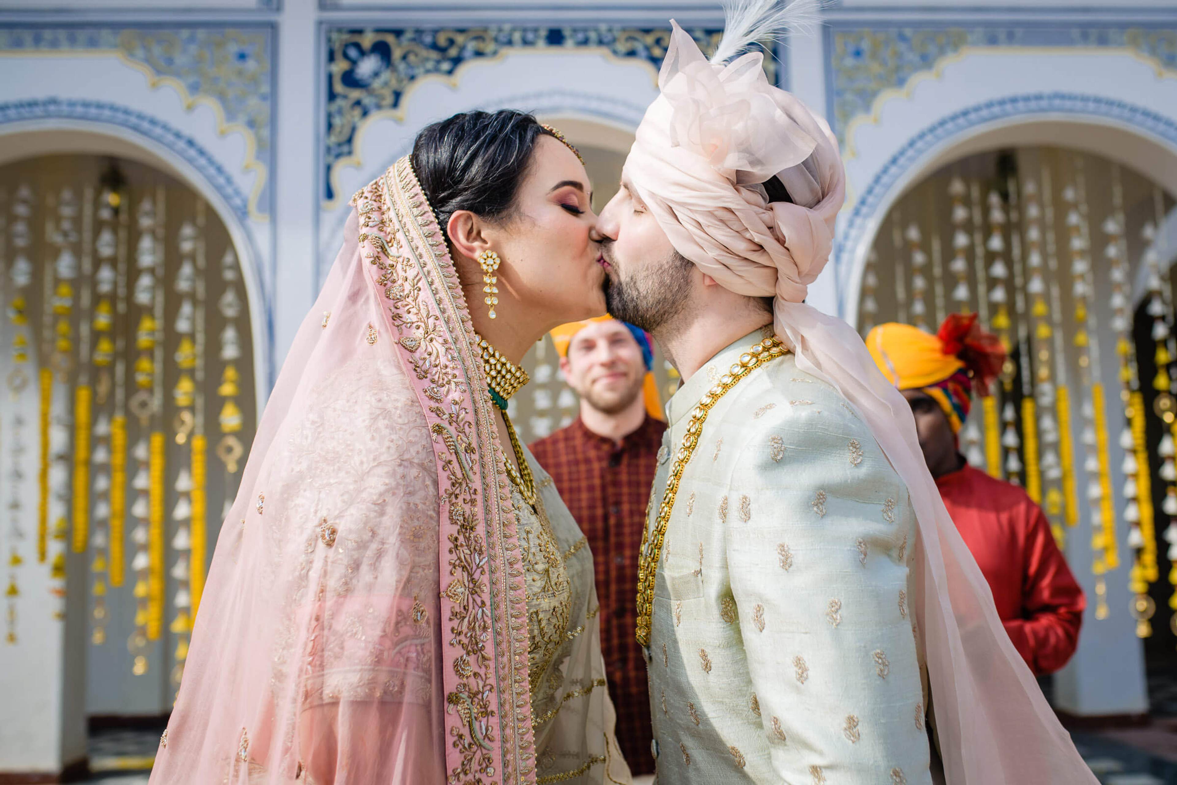 Maitrayee and Eric kiss in traditional South Asian wedding attire before ornate golden doors in Rajasthan