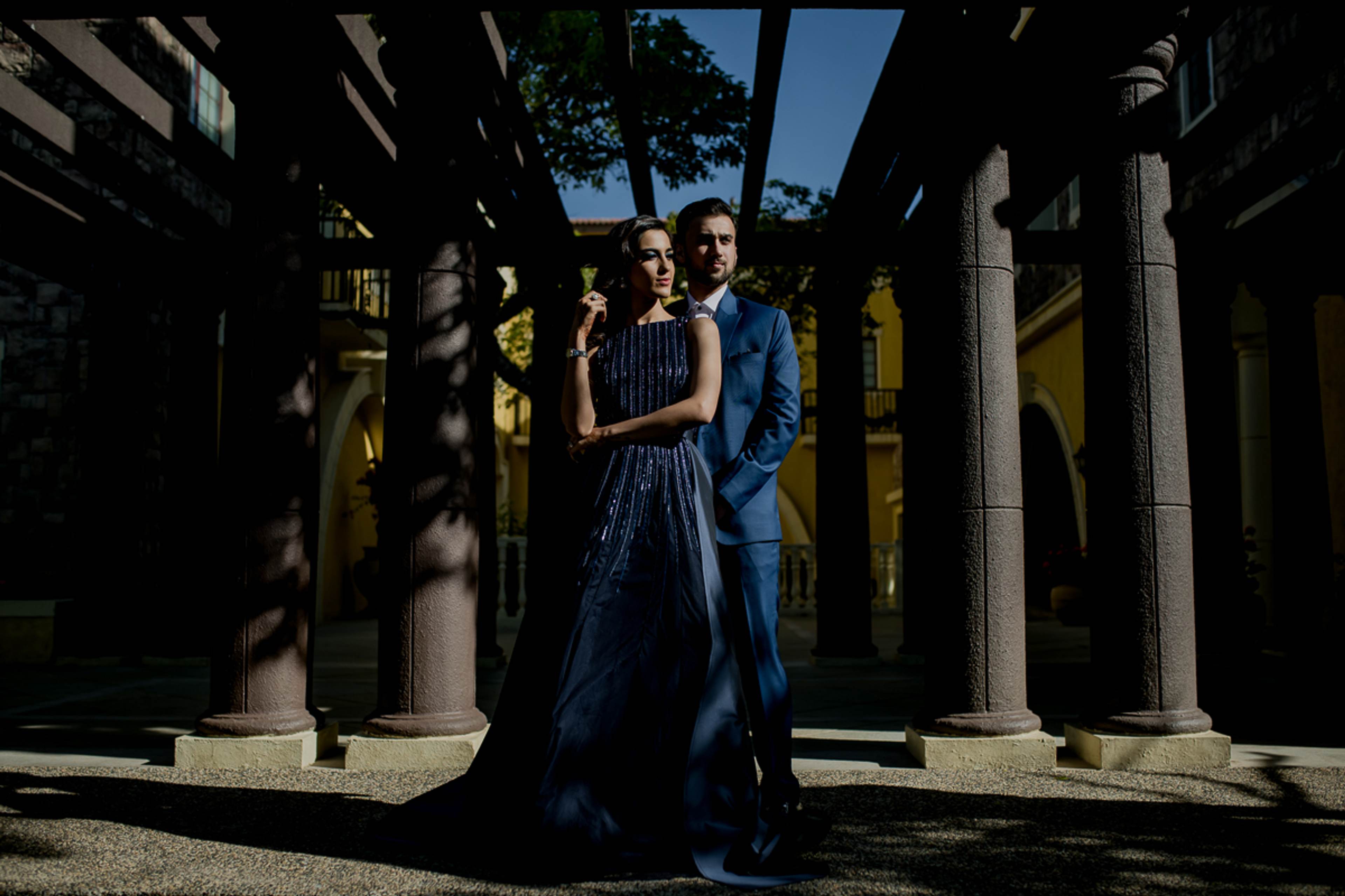 Couple in blue attire standing between columns with dramatic shadow and sunlight