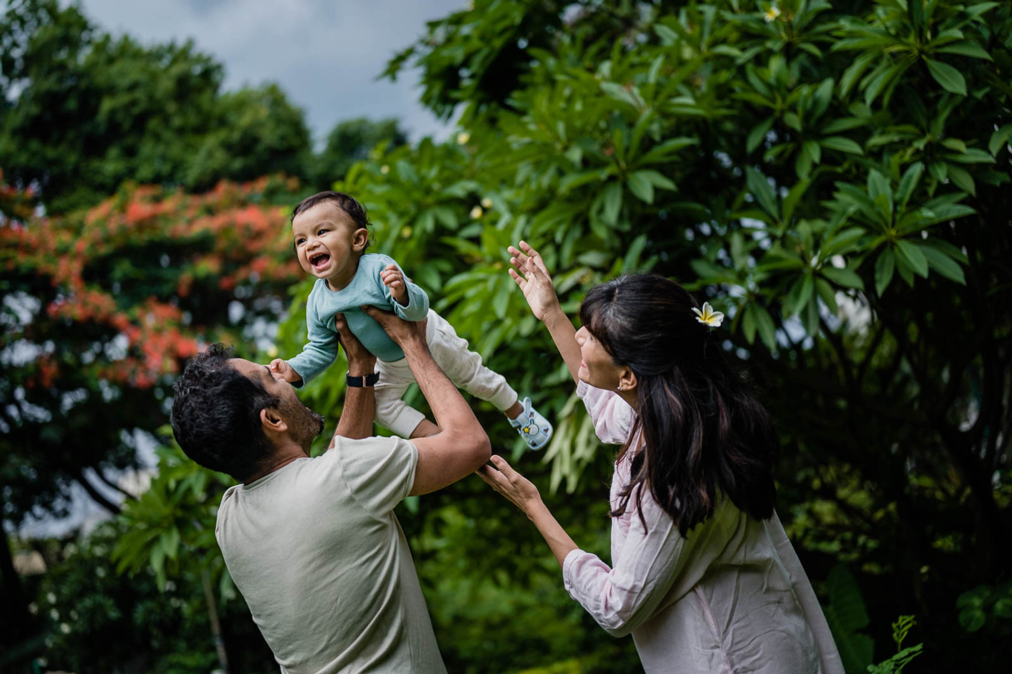 Man lifts young child as woman laughs, surrounded by lush green foliage and flowering trees
