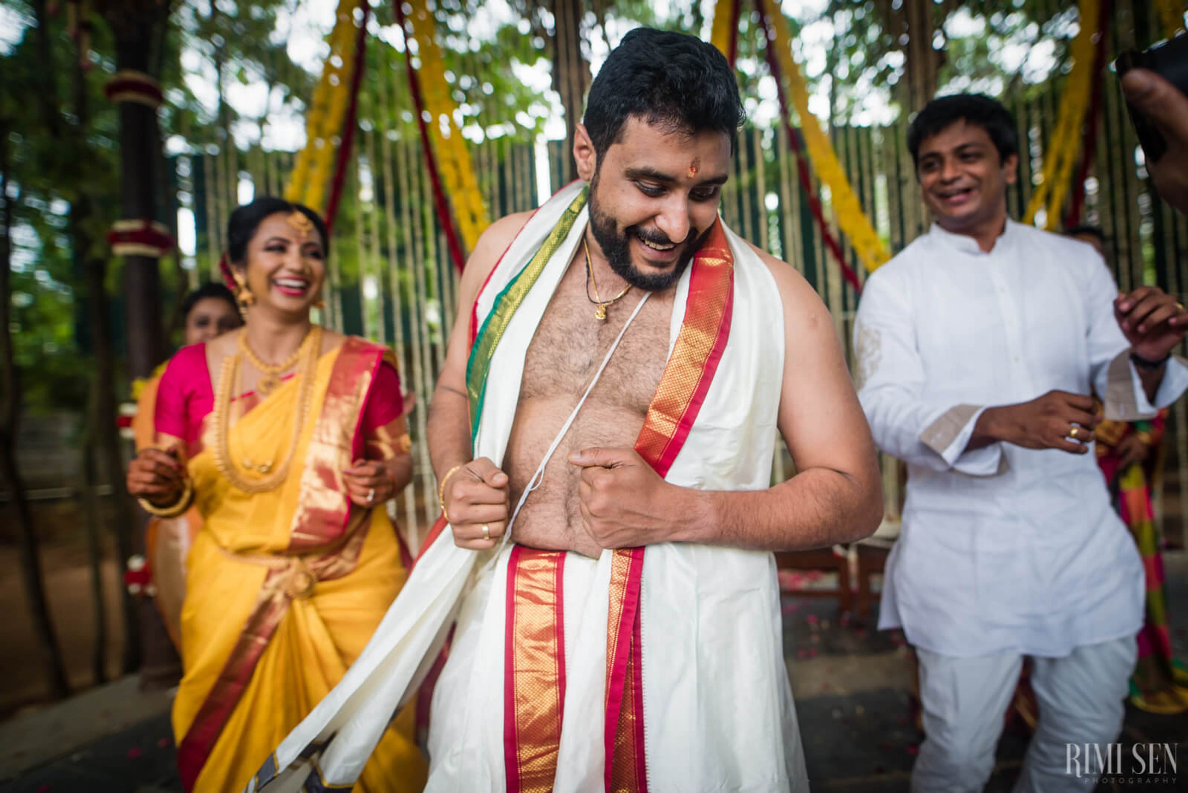 Groom Sridhar in traditional white and red silk draped attire, smiling during ceremony in bamboo pavilion, Bangalore