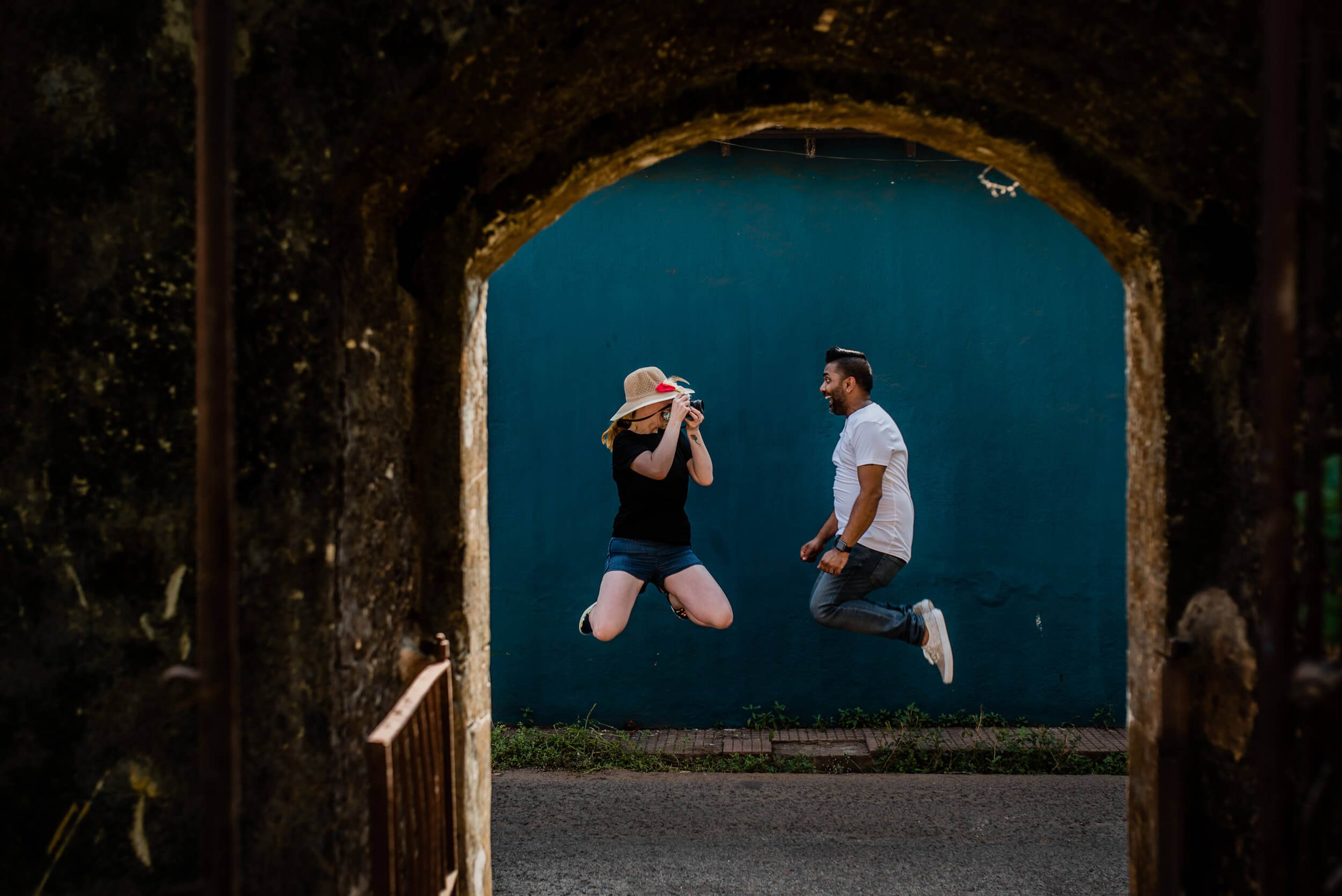 Sarah and Ravin jump joyfully against vibrant turquoise wall, framed by weathered stone archway in Goa
