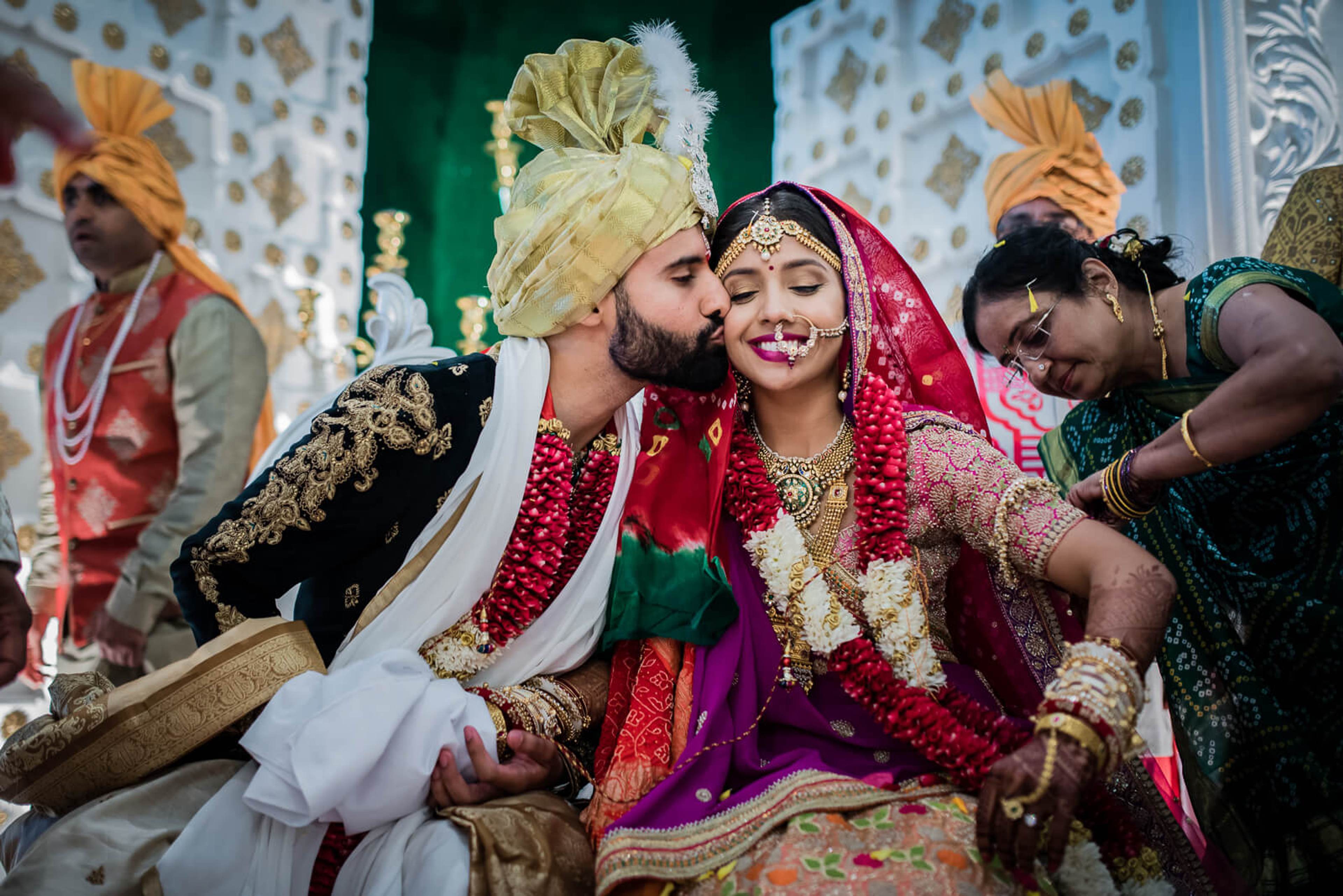 Groom in gold turban and embroidered sherwani kisses bride in red lehenga during Indian wedding ceremony