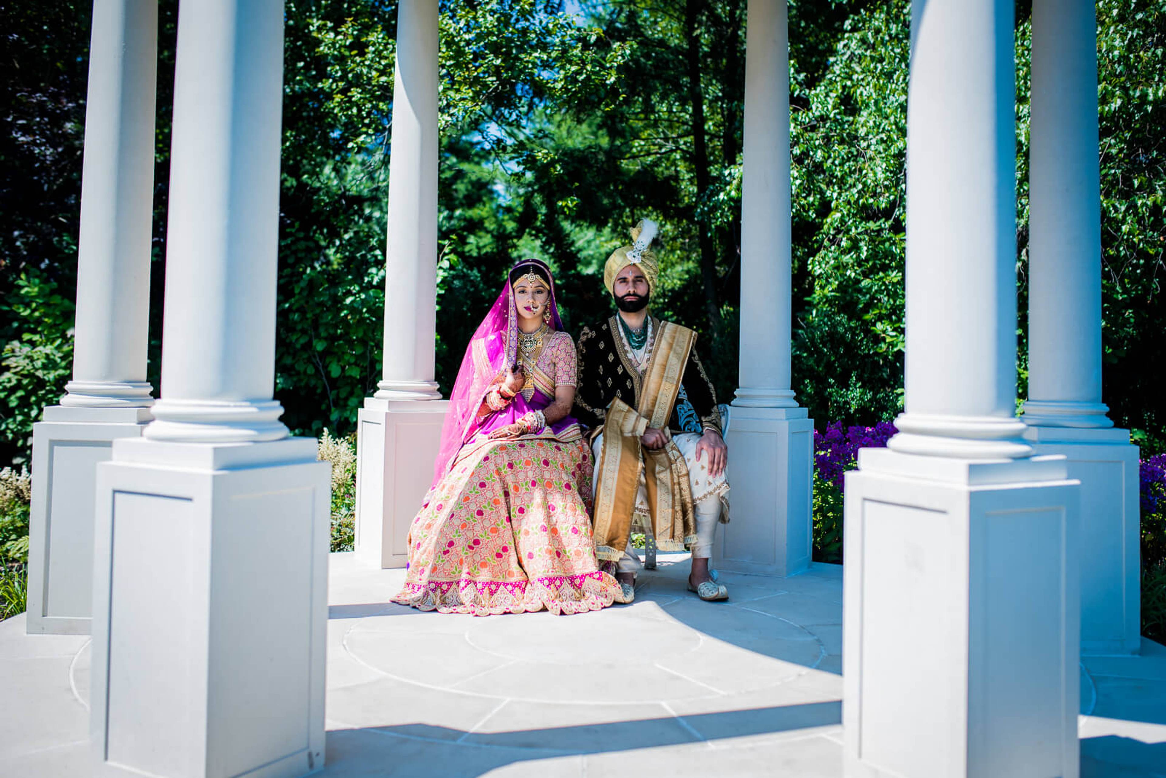 Barkha and Ankit sit together under ivy-covered columns in New York, wearing traditional Indian wedding attire