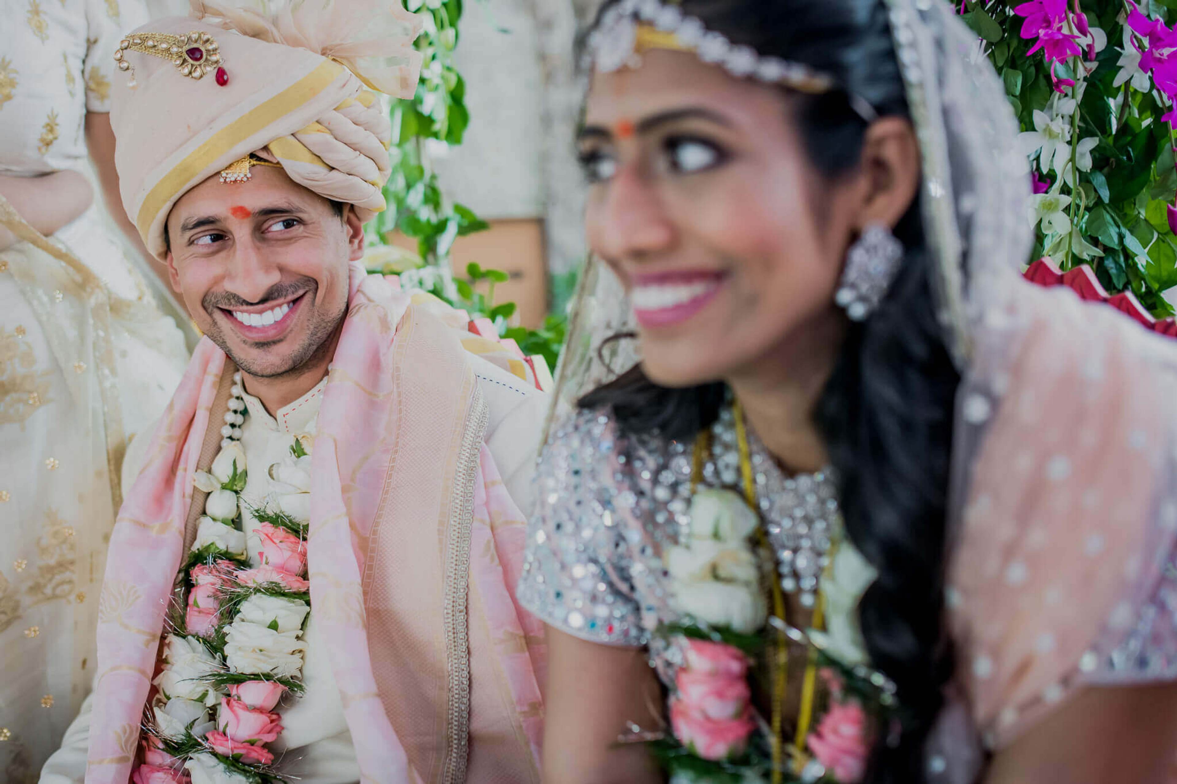 Groom Raghu in cream sherwani and pink turban smiles beside bride Anu in embroidered lehenga, vibrant flowers framing them
