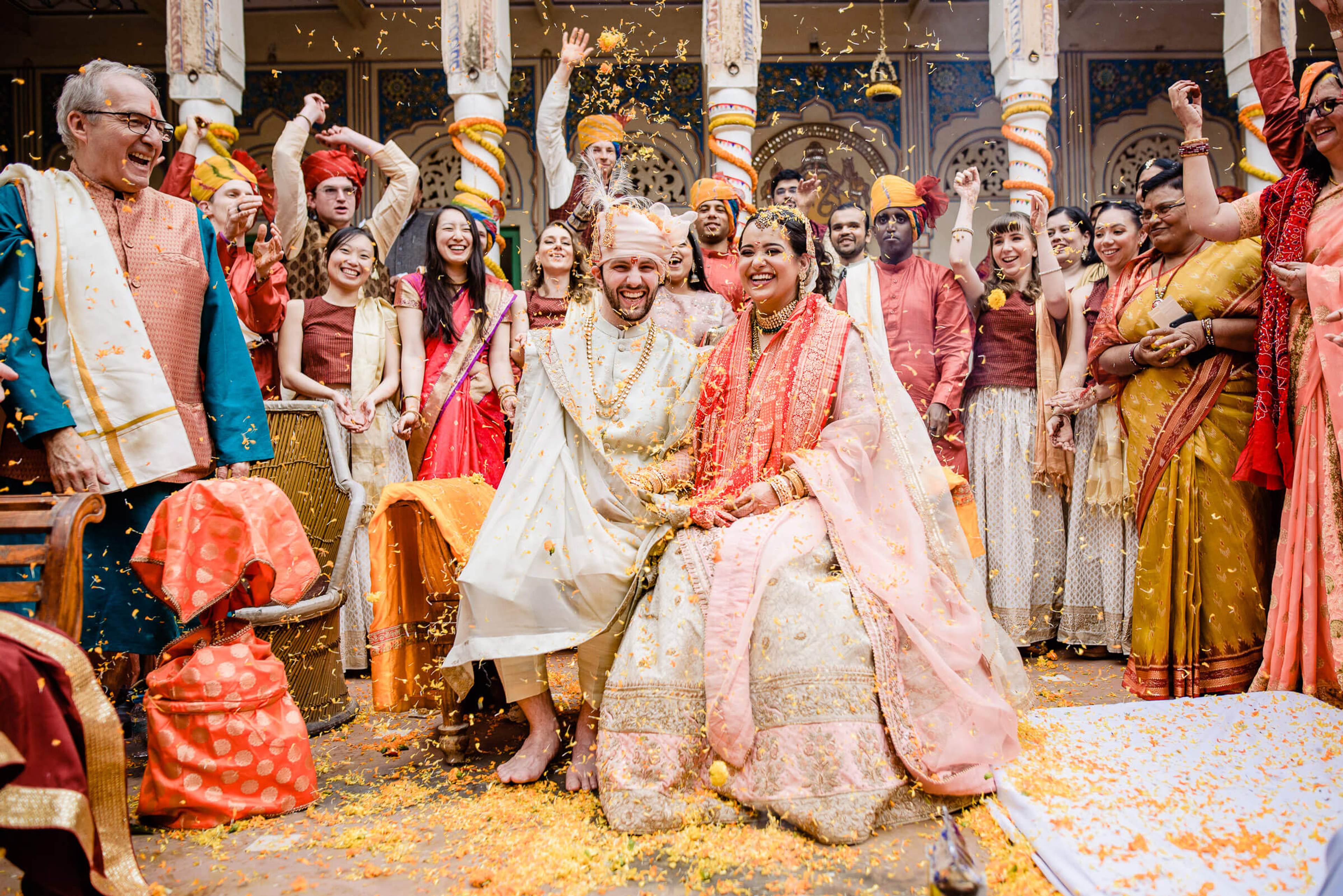 Maitrayee and Eric sit together in traditional wedding attire surrounded by celebrating guests throwing flower petals in Rajasthan