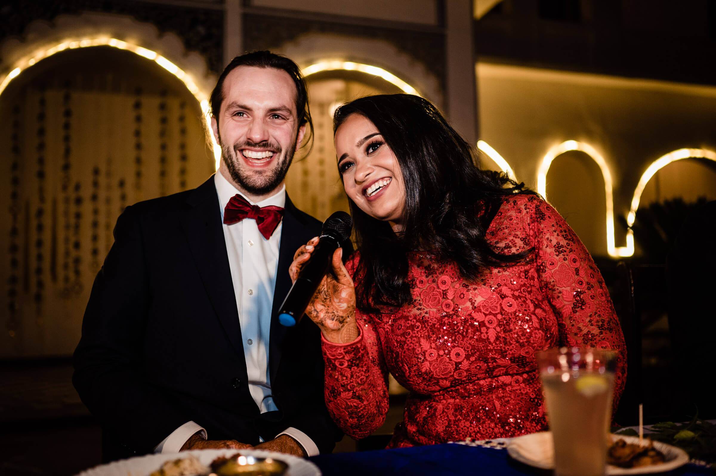 Maitrayee and Eric smile together at a candlelit table, he in a tuxedo and burgundy bow tie, she in a red sequined top