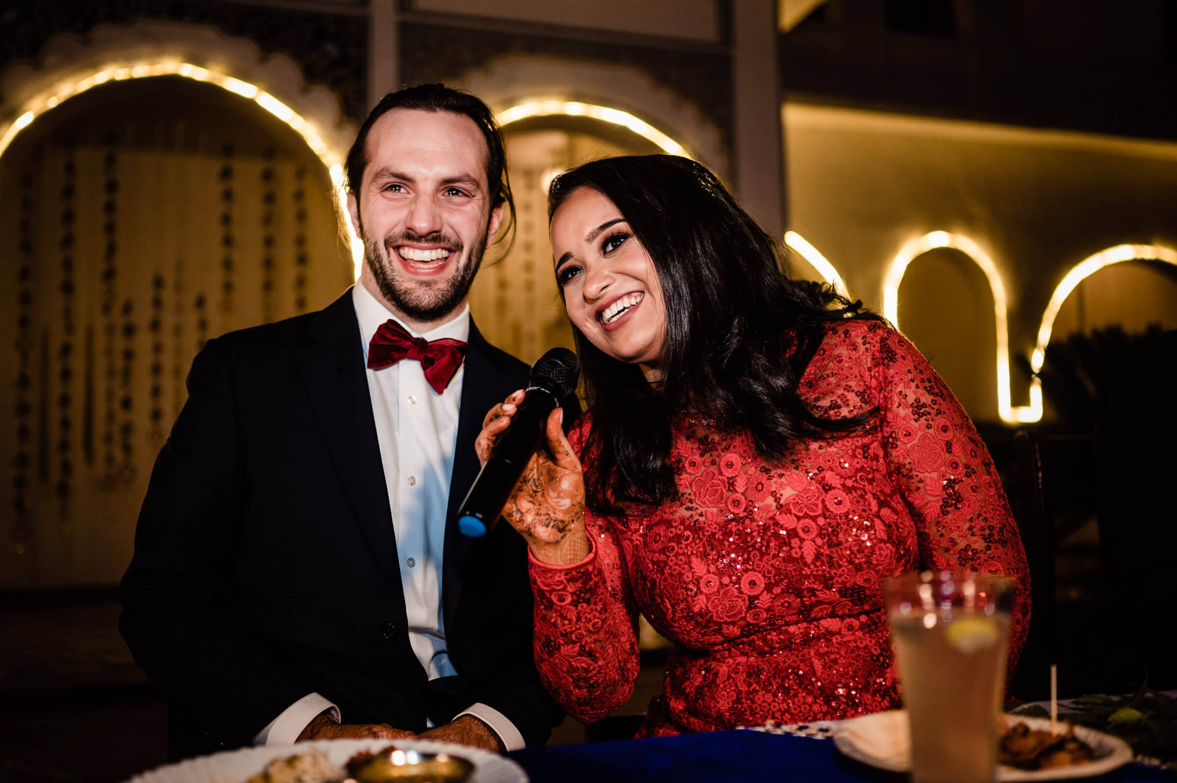 Maitrayee and Eric smile together at a candlelit table, he in a tuxedo and burgundy bow tie, she in a red sequined top