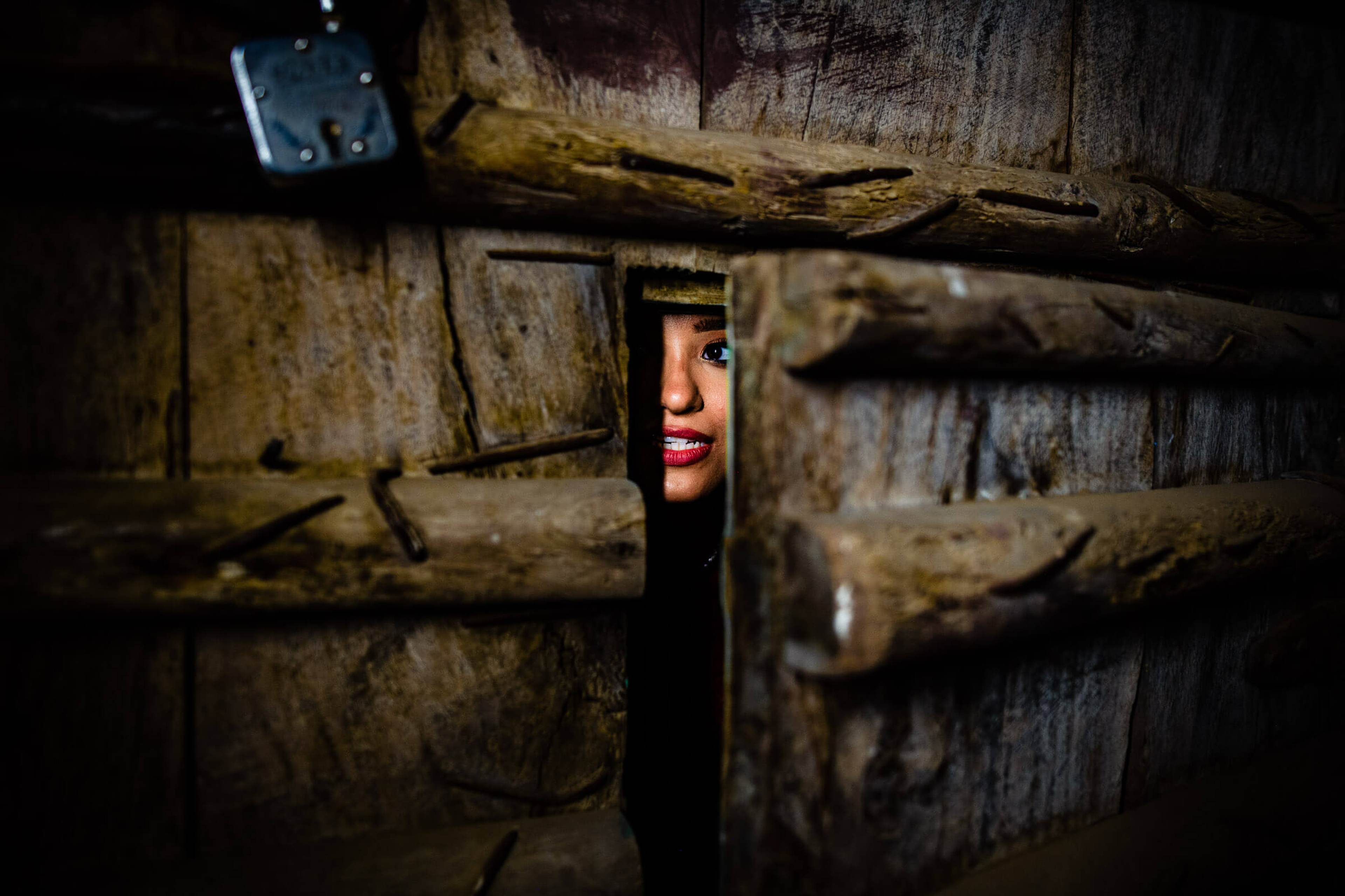 Maitrayee peeks through rustic wooden beams in Rajasthan, red lips visible amid weathered timber
