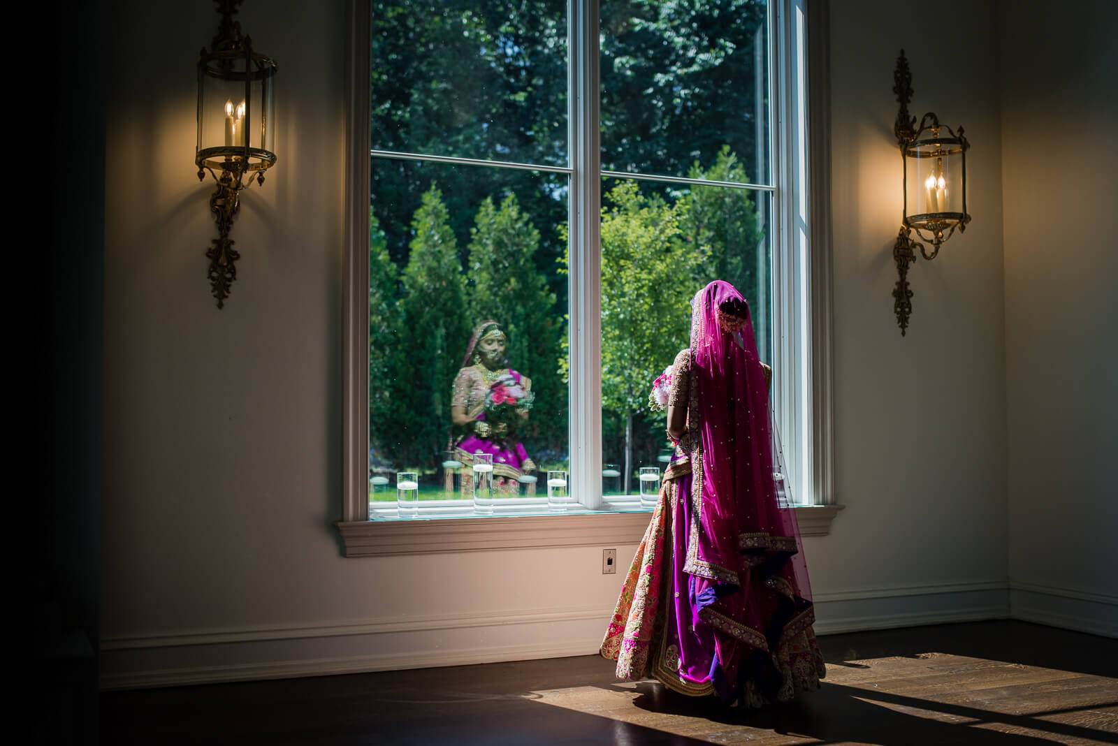 Barkha in vibrant magenta lehenga gazes through tall windows at garden reflection, ornate sconces flanking