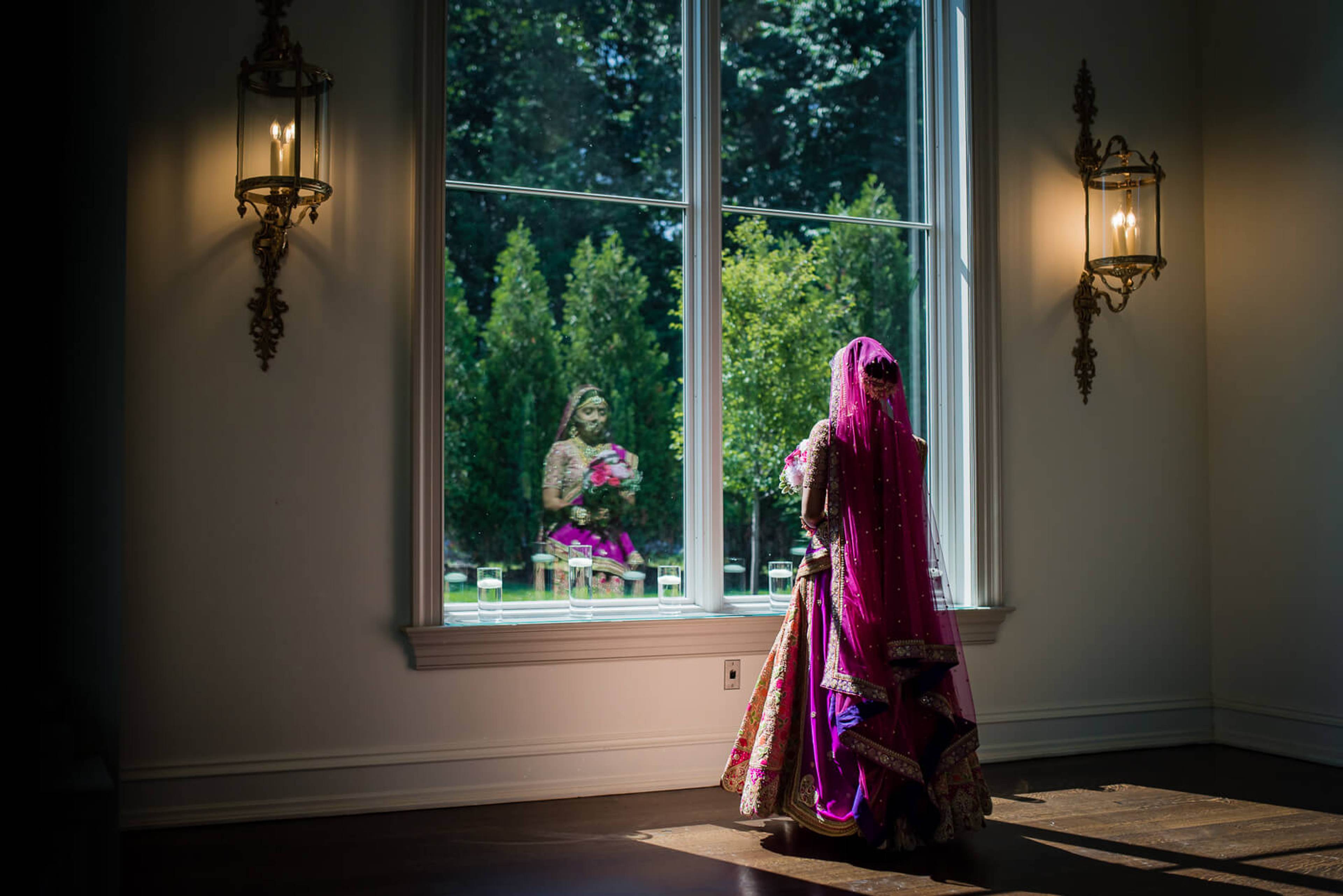 Barkha in vibrant magenta lehenga gazes through tall windows at garden reflection, ornate sconces flanking