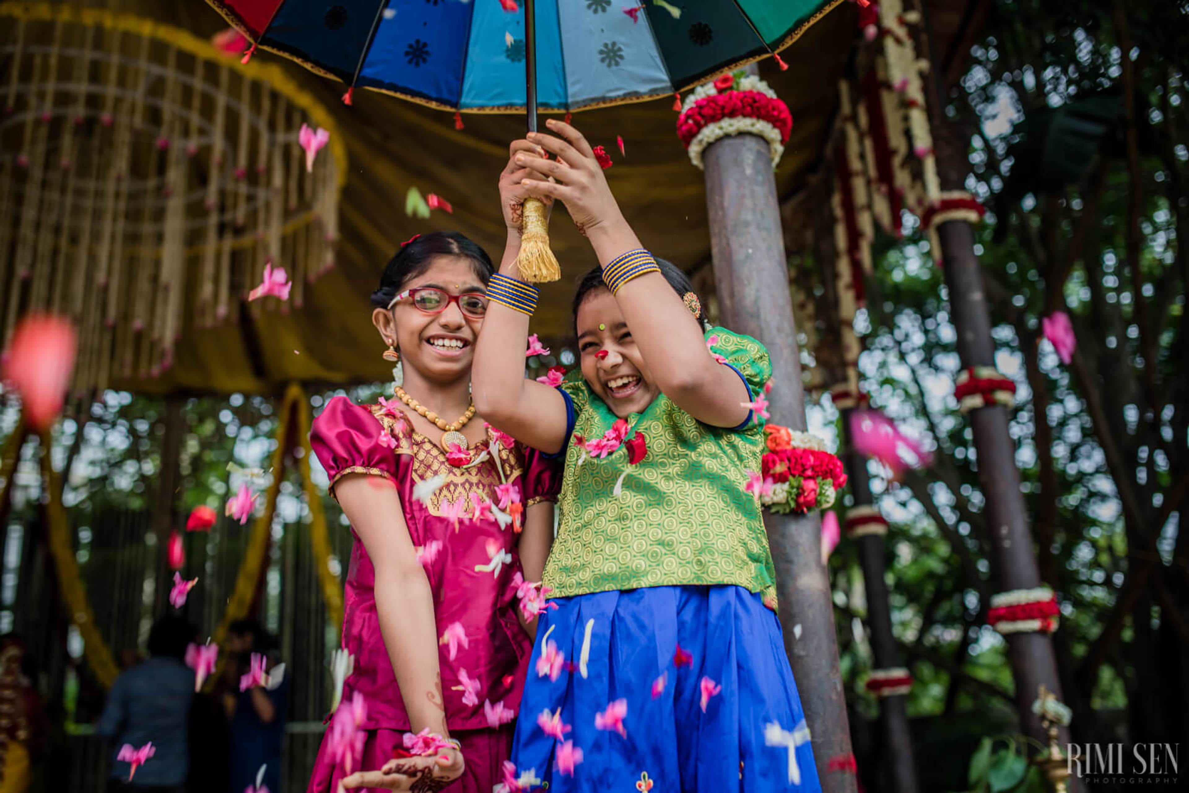 Two women in vibrant traditional attire holding colorful umbrella under flower petals in Bangalore