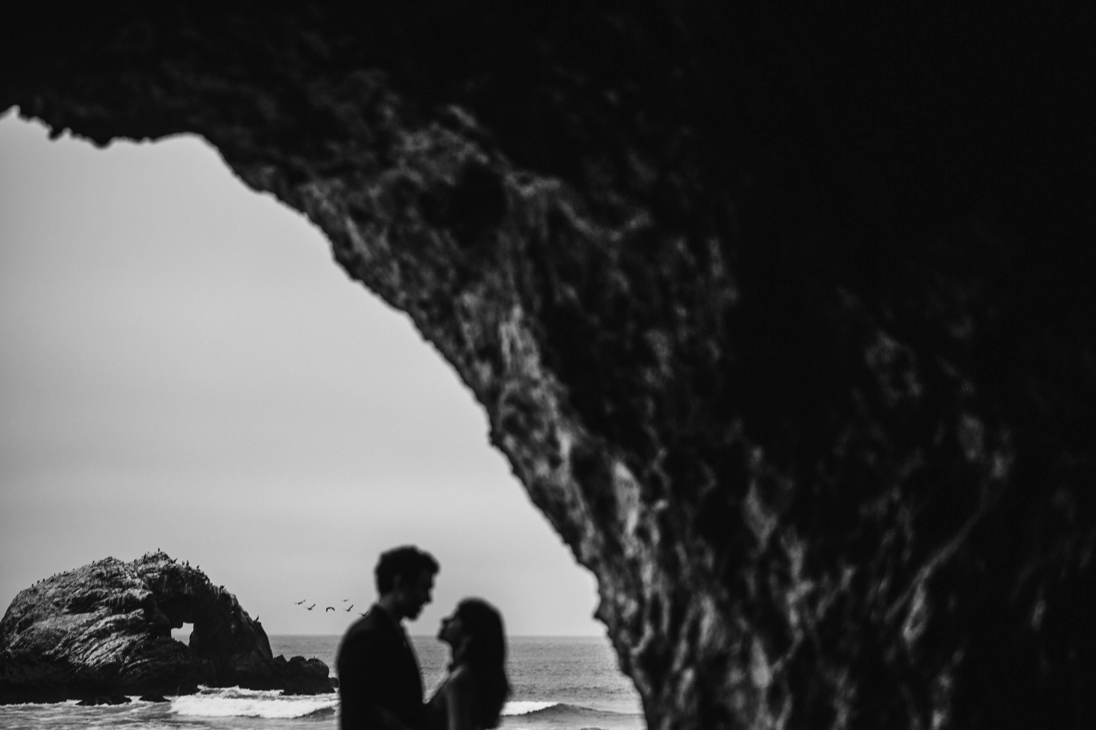 Couple framed through a rocky cave opening, standing on beach with ocean and rock formations behind, black and white