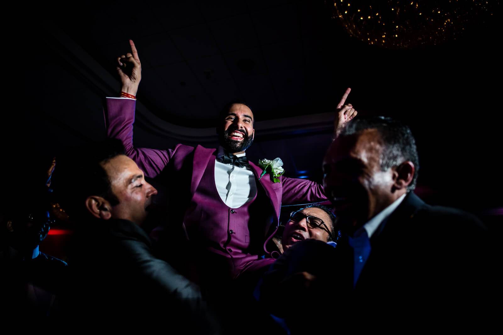 Groom in purple jacket celebrating with guests at New York wedding reception, arms raised, smiling