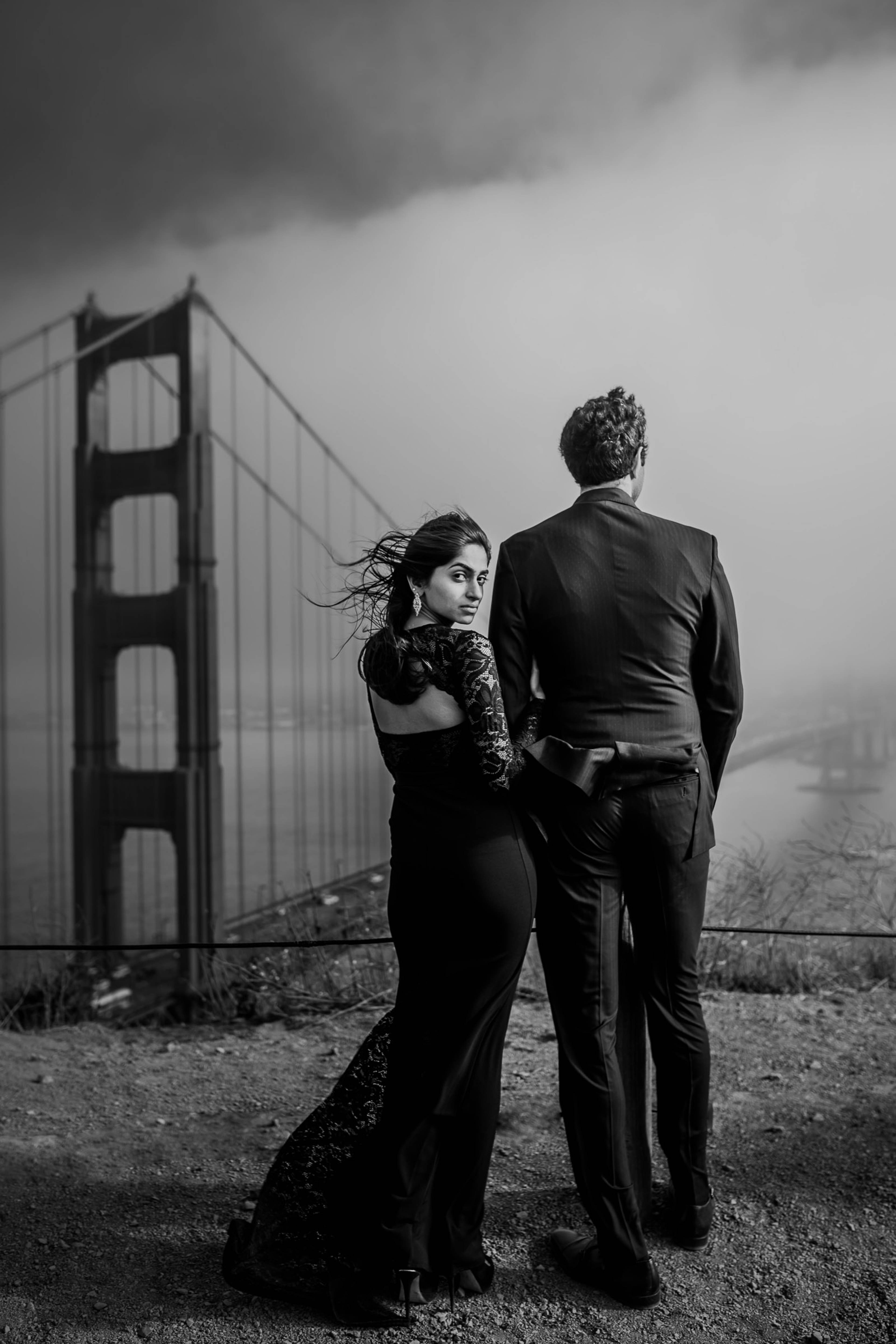 Couple in formal black attire standing before Golden Gate Bridge under dramatic cloudy sky, black and white portrait