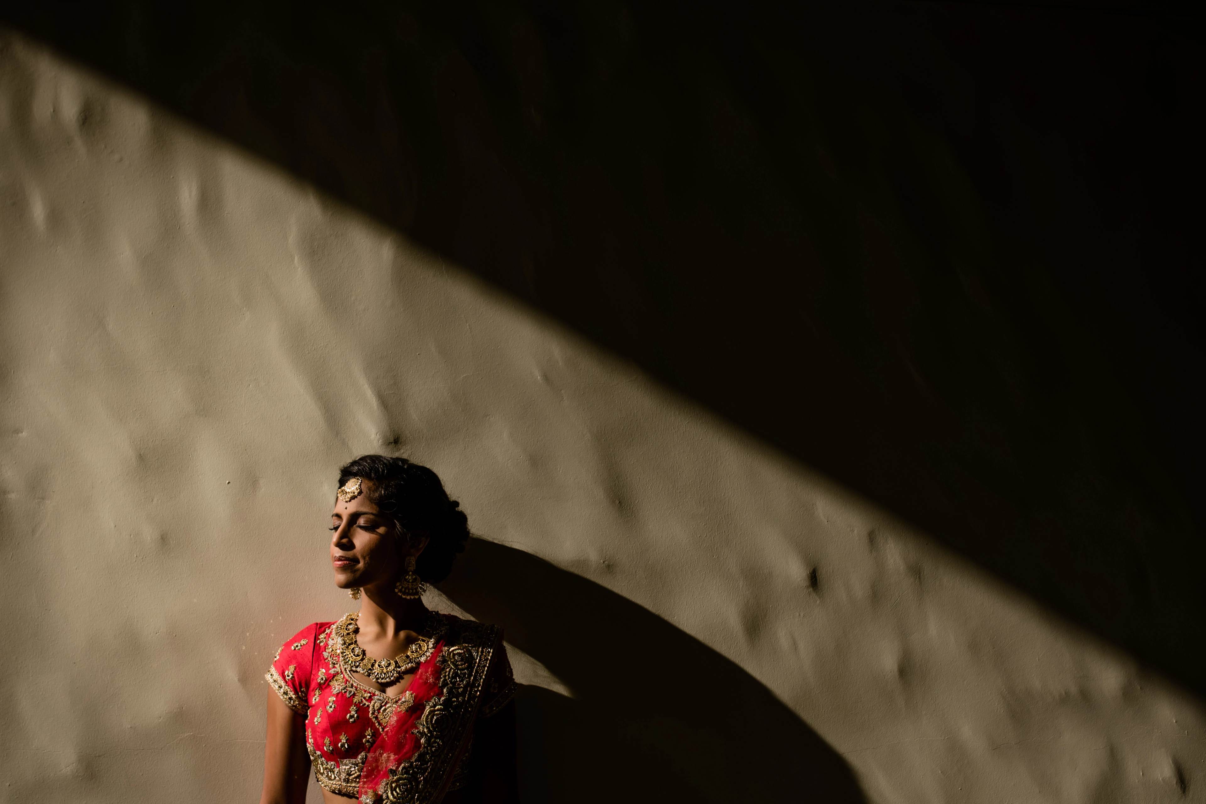Woman in red embroidered dress lit by dramatic sunlight against textured white wall