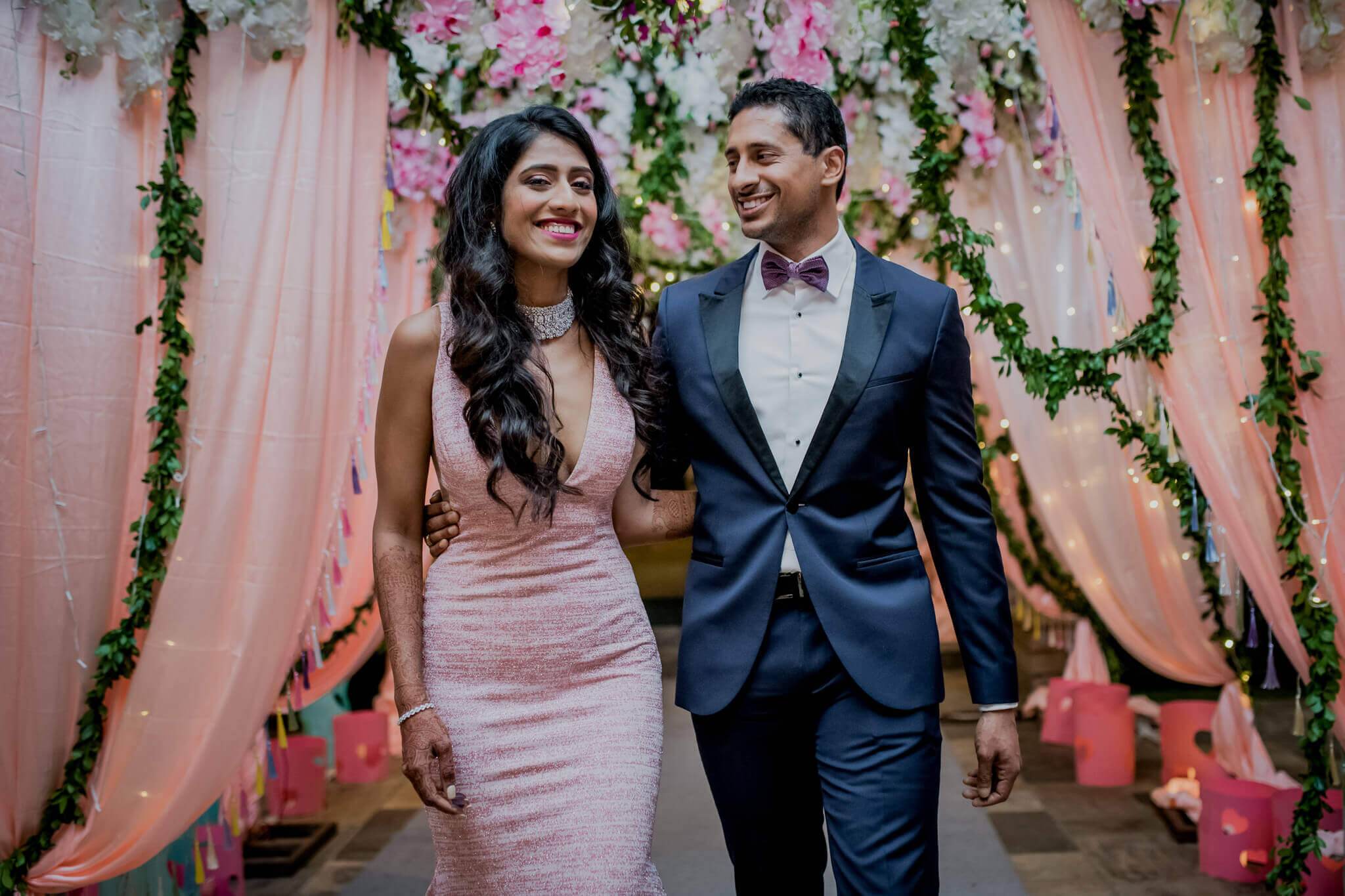 Anu and Raghu walk beneath pink draped fabric and floral garland at their Nepal wedding venue