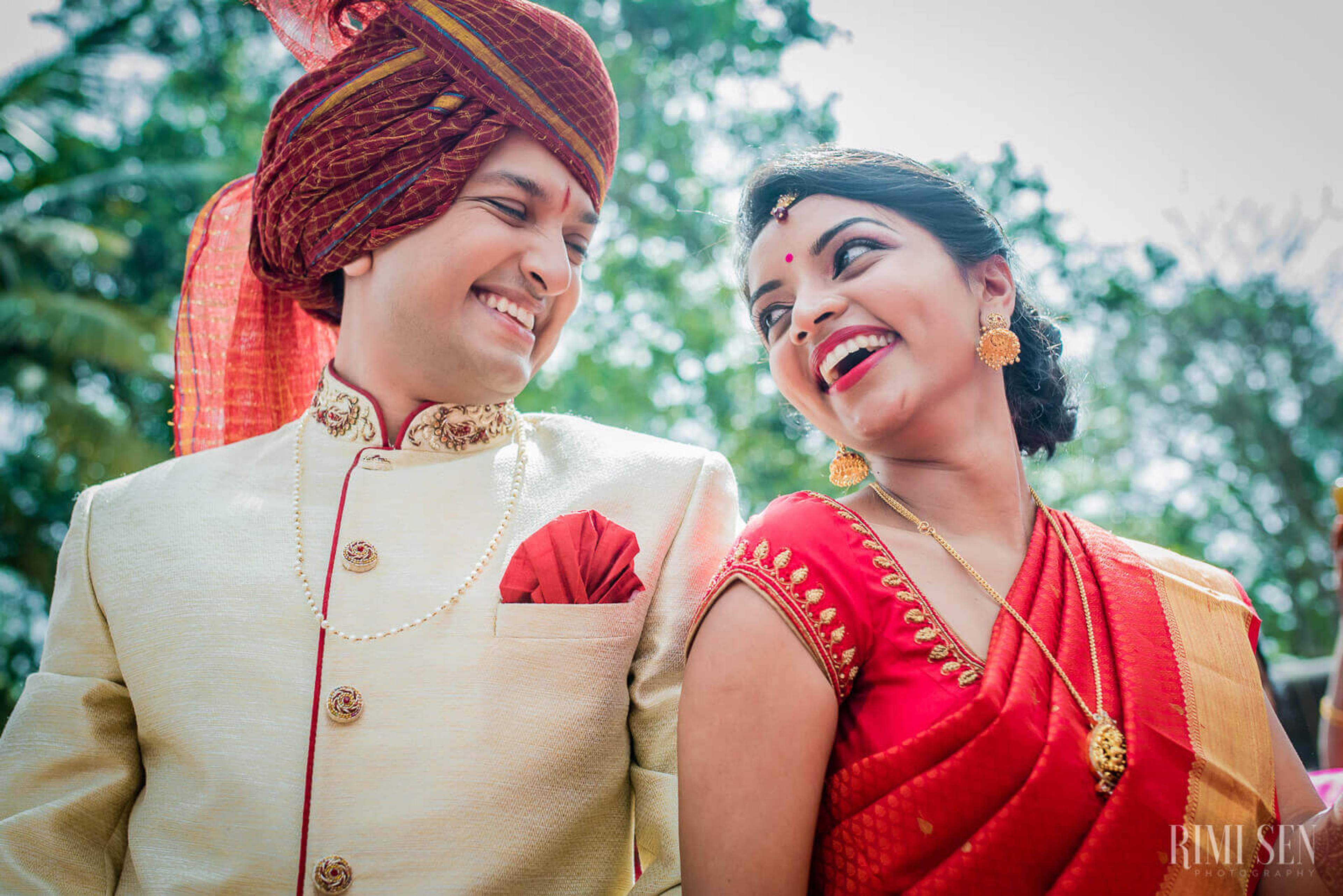 Pooja and Aniket smile together in traditional Indian wedding attire against lush green trees in Bangalore