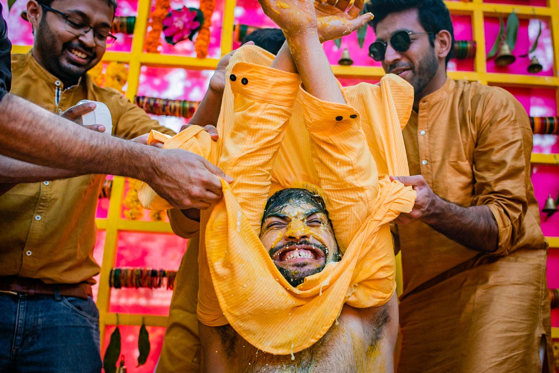 Two men in golden shirts holding up a yellow fabric with a painted face against bright pink and yellow backdrop in Goa