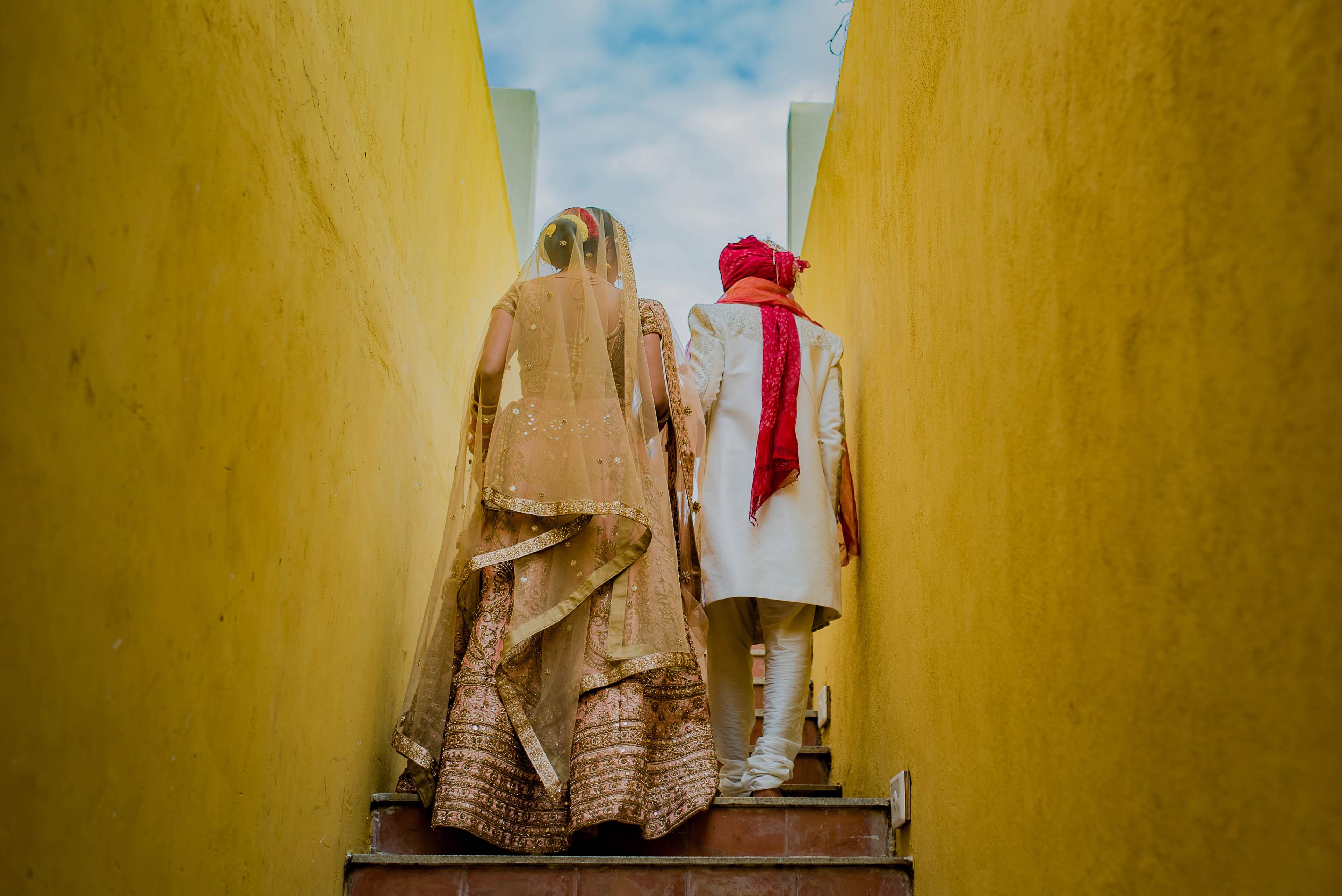 Couple walking through narrow yellow passage, bride in gold lehenga, groom in white sherwani with red scarf