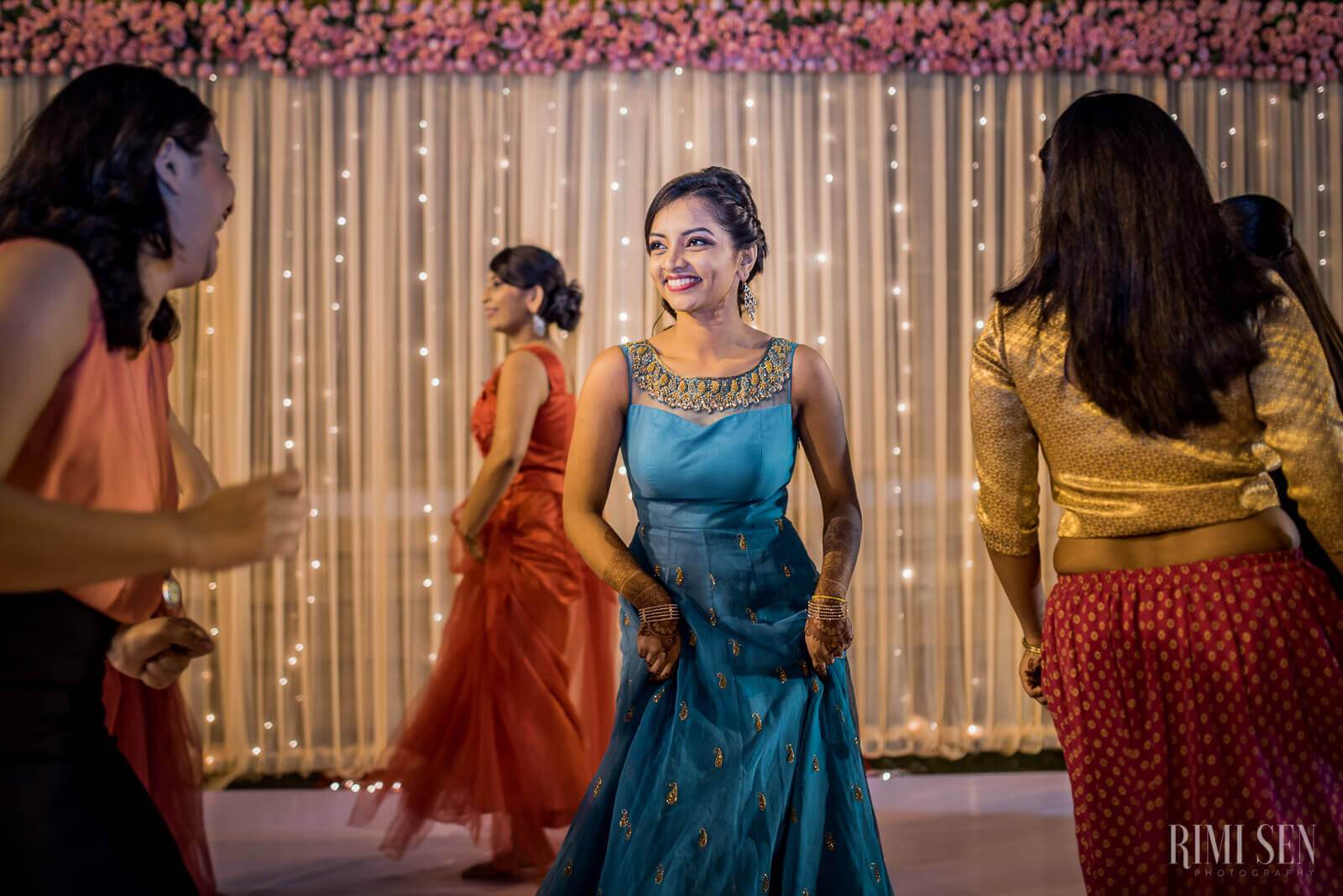 Woman in blue embellished dress smiles among guests in vibrant festive attire and twinkling lights backdrop
