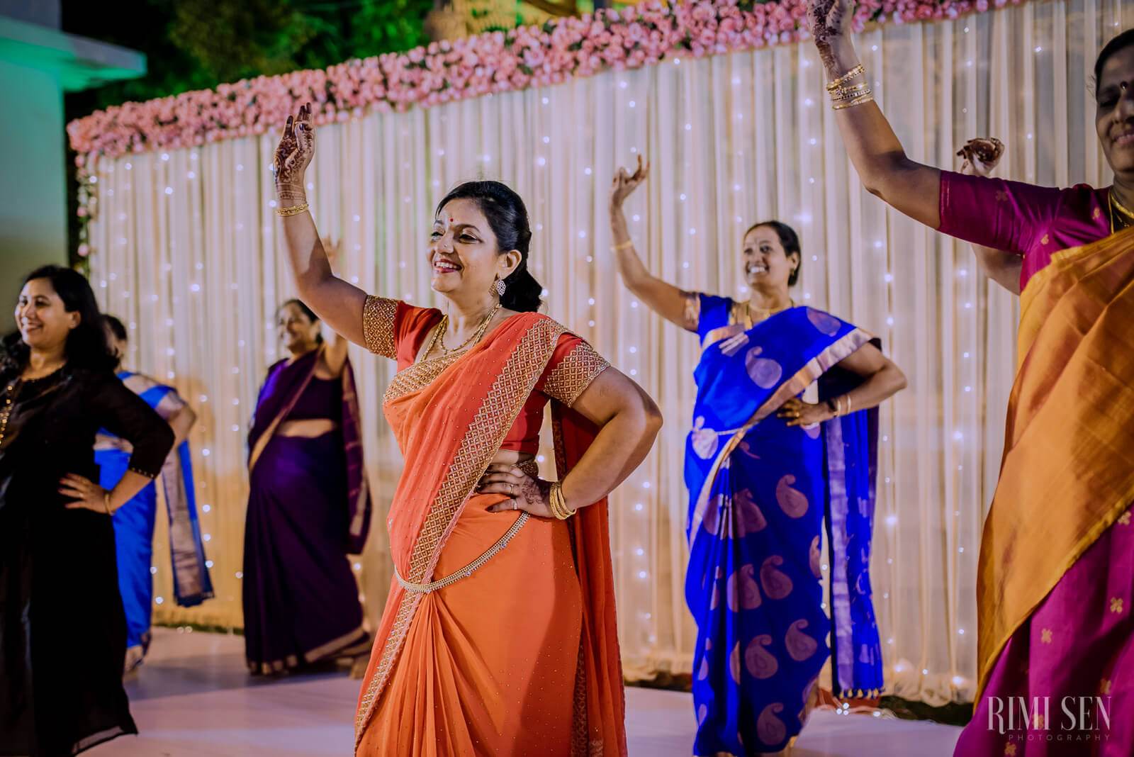 Woman in orange saree dancing with arms raised at Bangalore wedding celebration, pink floral backdrop